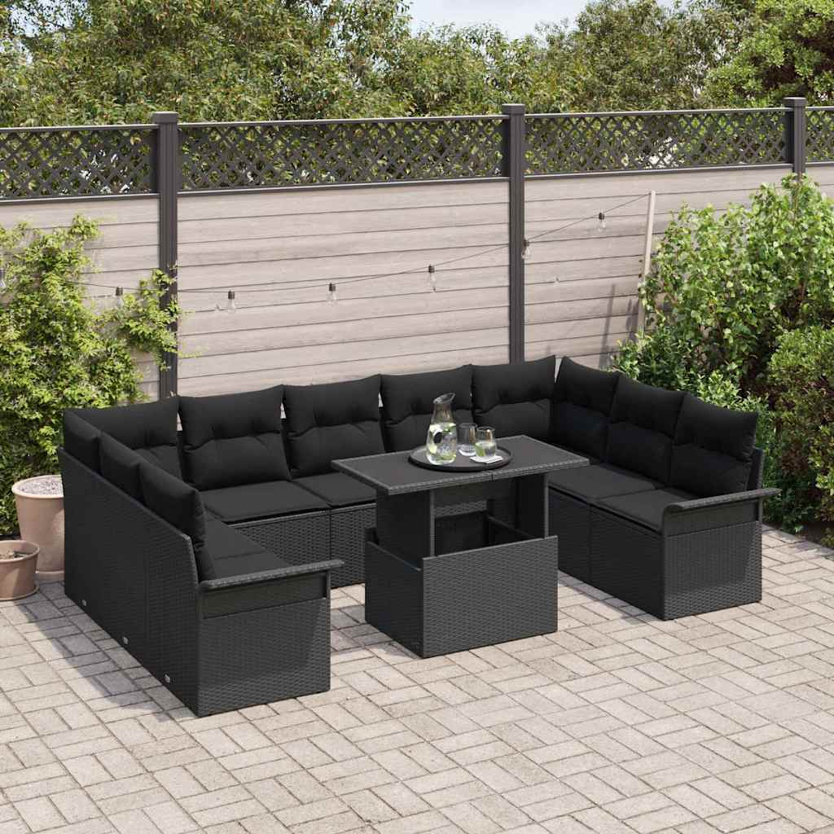 Ein schwarzes Outdoor-Ecksofa mit Tisch, Wasserkrug und Gläsern steht auf einer Terrasse. Grüne Büsche und ein Zaun sind im Hintergrund.