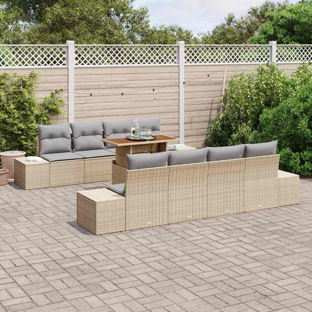 Juego de asientos al aire libre en un patio. Muebles de mimbre con cojines grises incluyen sofá, mesa y otomanas. Valla de madera y vegetación.