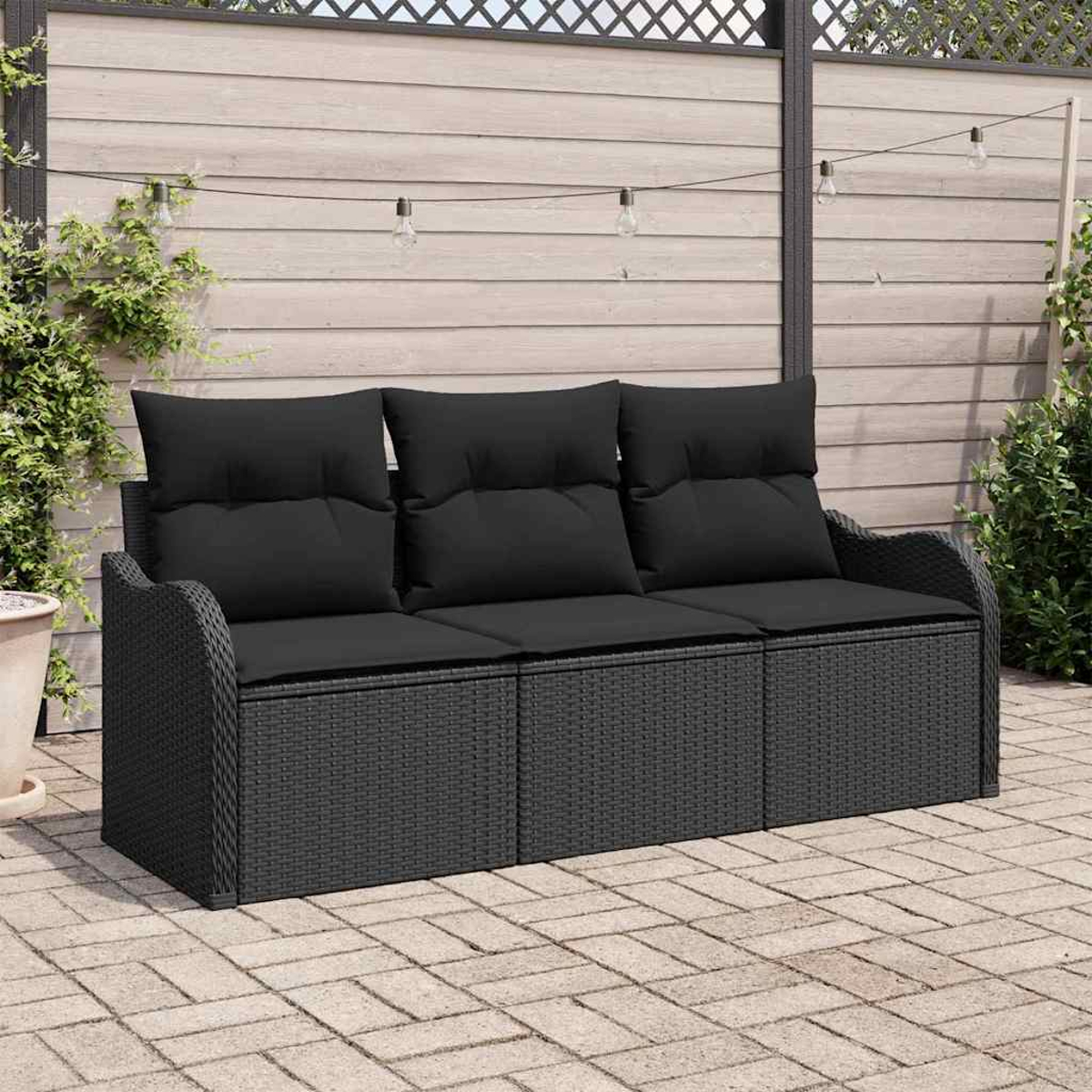 Ein schwarzes Outdoor-Sofa aus Korbgeflecht mit schwarzen Kissen steht auf einer Ziegelterrasse. Im Hintergrund sind ein Holzzaun und Lichterketten.