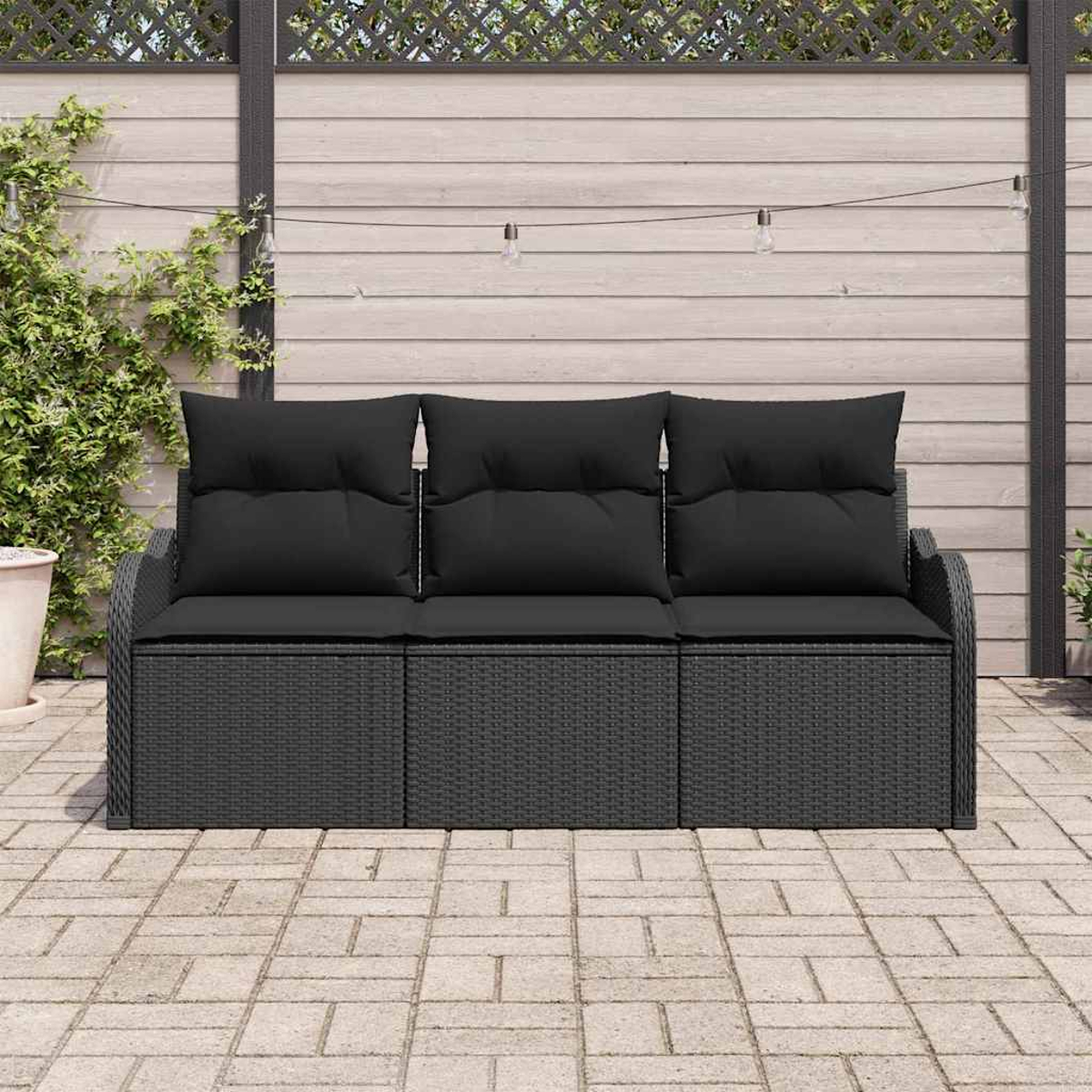 Schwarzes Korbmöbelsofa mit schwarzen Kissen auf einer Terrasse. Holzzaun und Lichterketten sind im Hintergrund. Grüne Pflanze links.