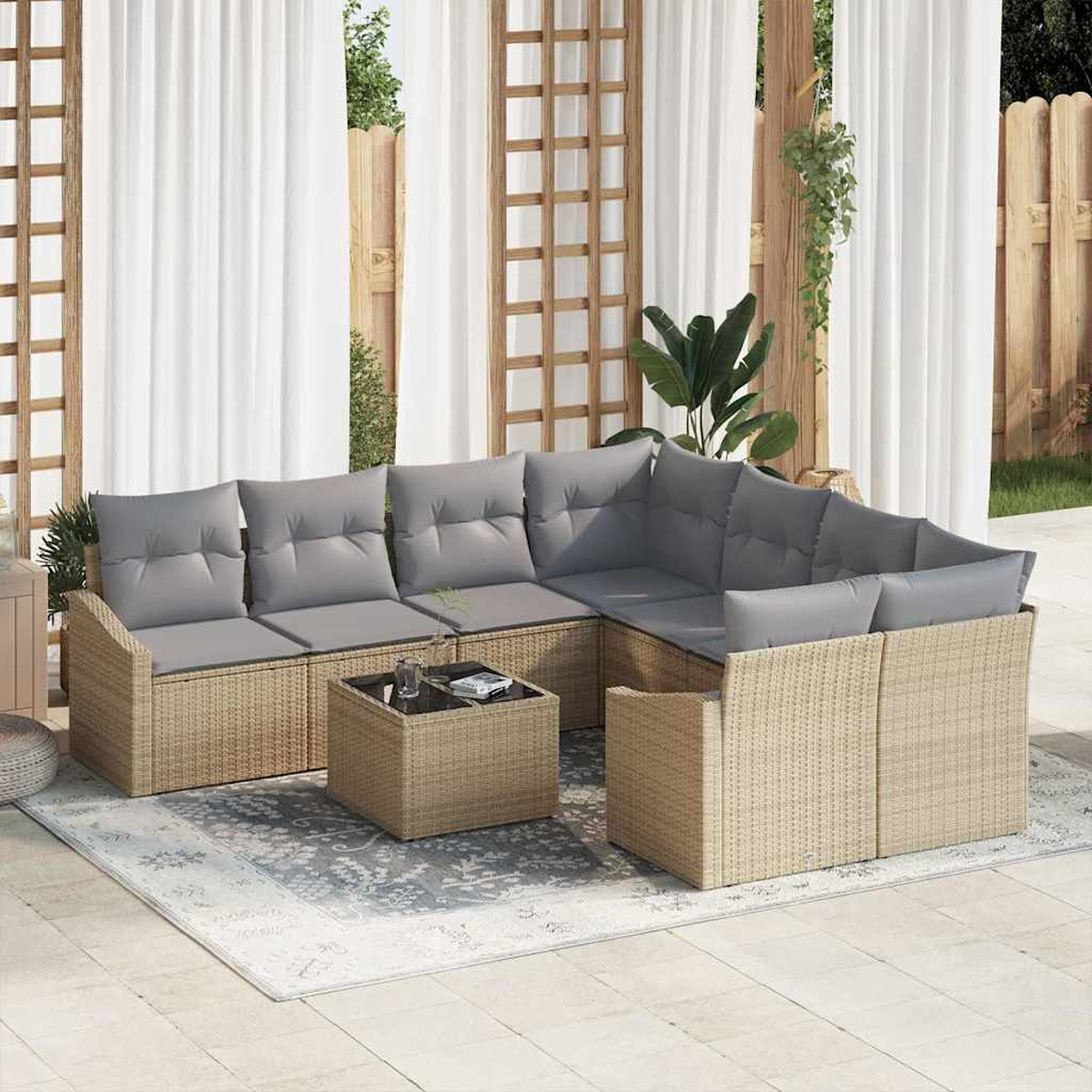 Sofá seccional de ratán para exteriores con cojines grises, mesa de centro y alfombra. Fondo con cortinas, marcos de madera y plantas.