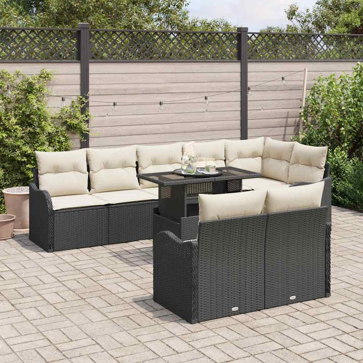 Ein schwarzes Outdoor-Ecksofa aus Korbgeflecht mit beigen Kissen und einem Tisch. Die Szene spielt in einem Hinterhof mit Holzzaun.