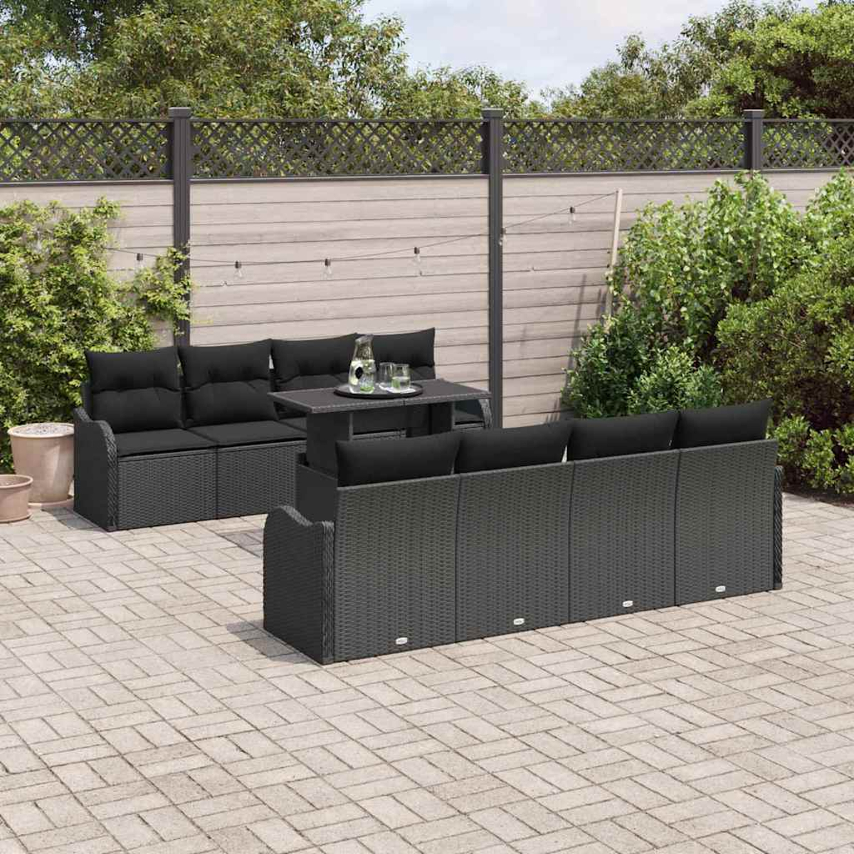 Terrassenset mit schwarzen Kissen auf grauen Korbmöbeln. Tisch mit Wasser und Gläsern. Zaun und Grün im Hintergrund.