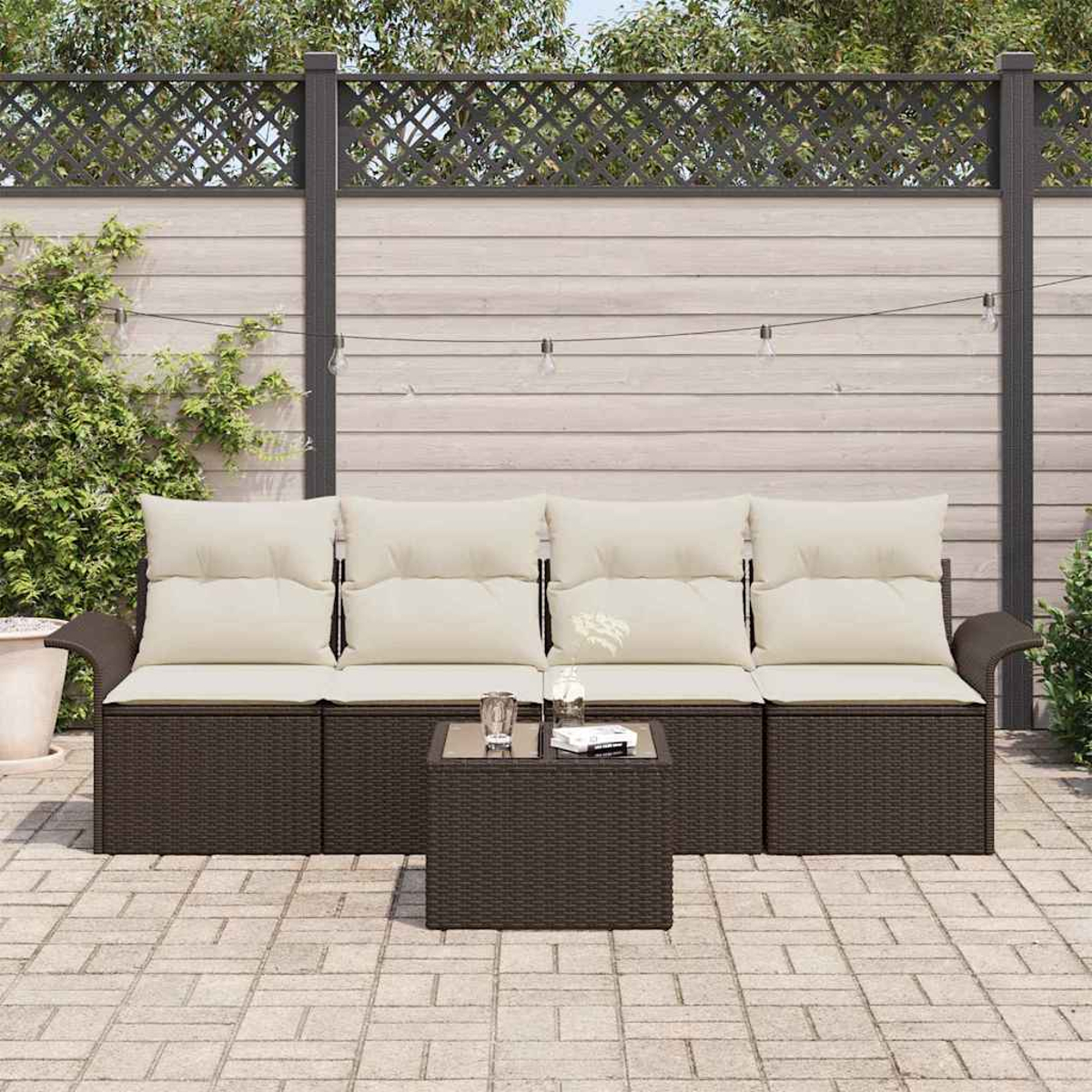 Terrassenset mit braunen Korbmöbeln, cremefarbenen Kissen und einem Tisch mit Glasplatte. Im Hintergrund ein Holzzaun und Pflanzen.