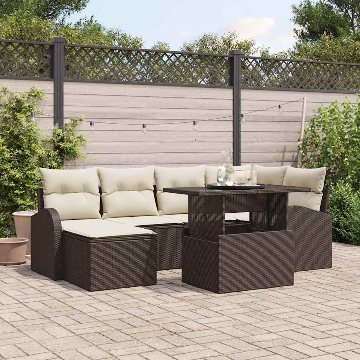 Terrassenmöbel-Set mit braunen Korbmöbeln, beigen Kissen und einem Tisch. Der Hintergrund zeigt einen Zaun und Grün.