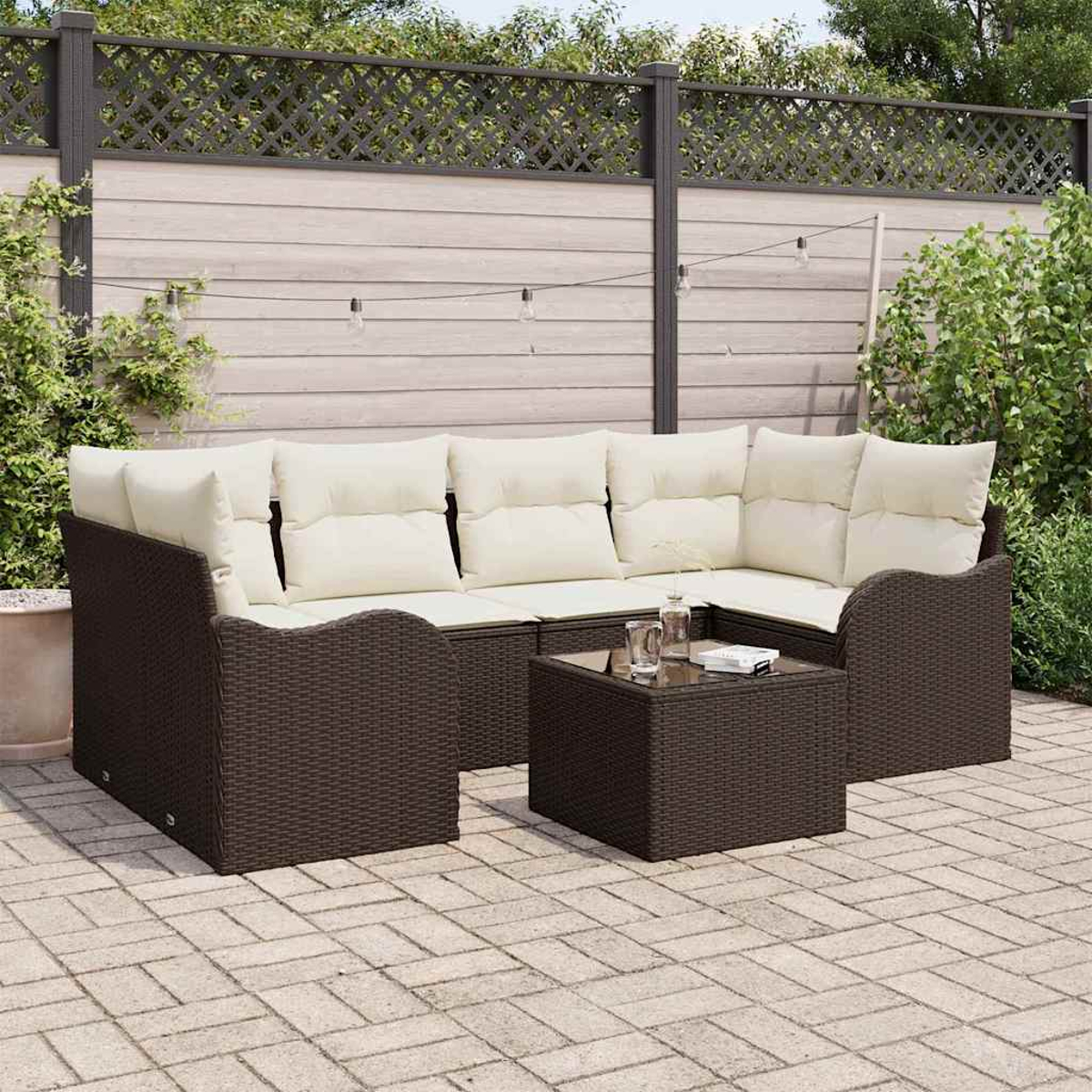 Outdoor-Sitzgruppe mit braunem Korbgeflecht, cremefarbenen Kissen und einem kleinen quadratischen Tisch. Dahinter ein Holzzaun, Terrasse und Grün.