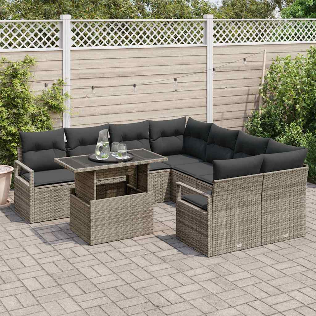 Garten-Sitzgruppe. Graue Korbmöbel mit schwarzen Kissen und einem Tisch mit Glasplatte. Auf einer Steinterrasse, im Hintergrund ein Zaun.