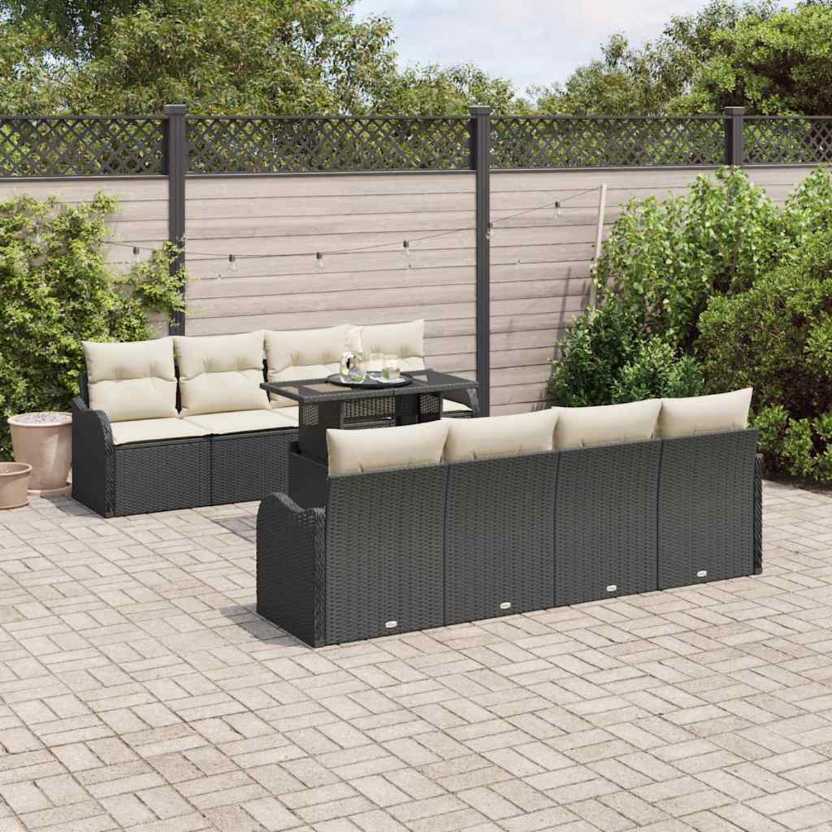 Terrassenmöbel-Set mit schwarzen Korbsesseln, beigen Kissen und einem kleinen Tisch auf einer gepflasterten Terrasse. Hintergrund: Holzzaun.