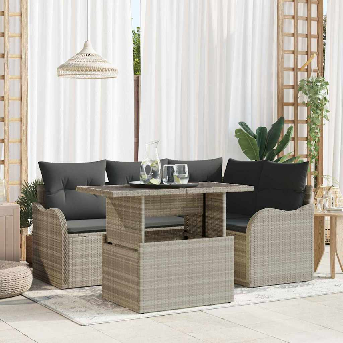 Gartenmöbel-Set aus Korbgeflecht mit grauen Kissen und Glaskrug auf einem Tisch vor weißen Vorhängen.
