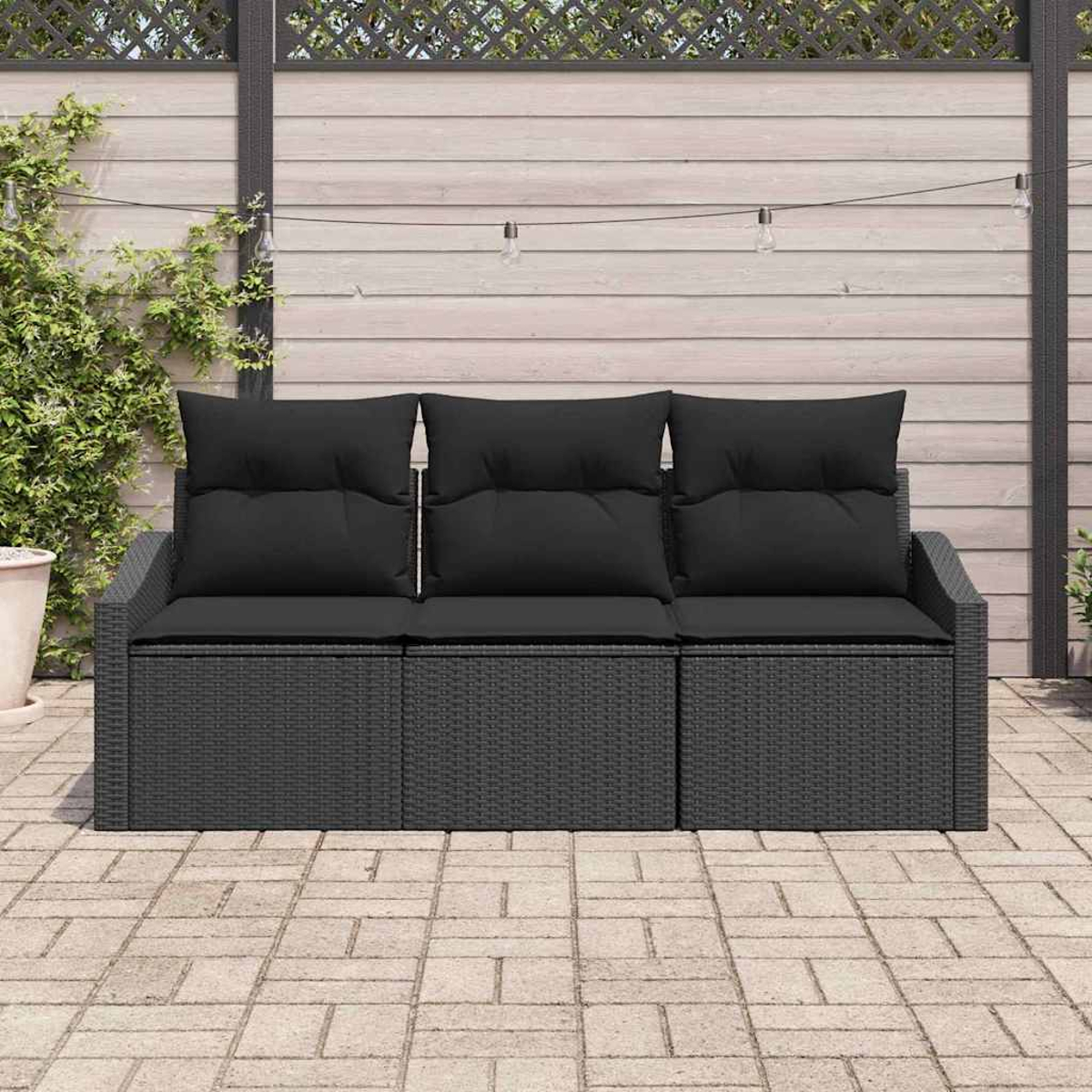 Ein dunkelgraues Rattan-Sofa mit schwarzen Kissen steht auf einer Ziegelterrasse. Im Hintergrund sind ein Holzzaun und Hängelampen zu sehen.
