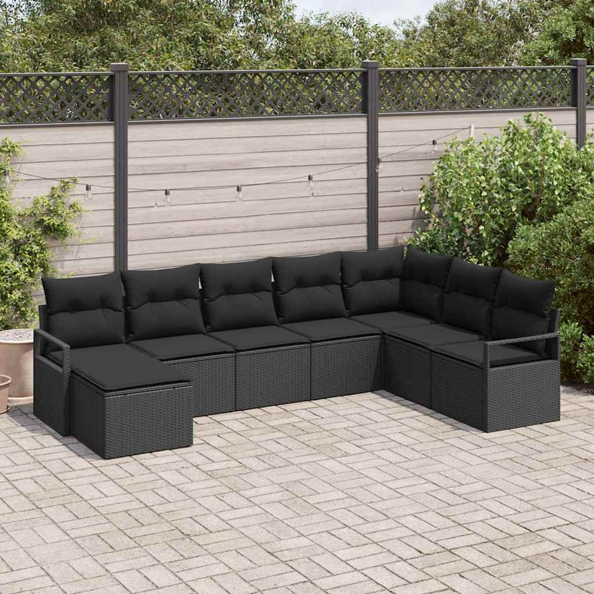 Ein schwarzes Ecksofa aus Korbgeflecht mit schwarzen Kissen steht auf einer Ziegelstein-Terrasse. Im Hintergrund sind ein Holzzaun und Grün.