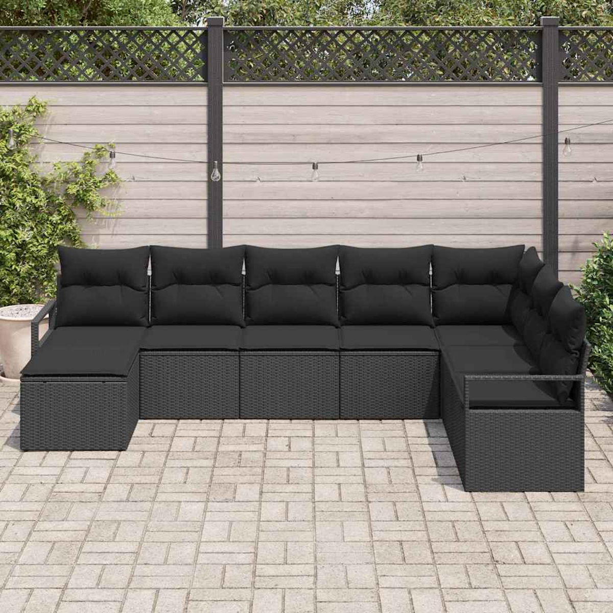 Schwarzes Rattan-Ecksofa auf einer Terrasse. Im Hintergrund ein Holzzaun und Lichterketten. Grüne Büsche und Pflanzen an der Seite.