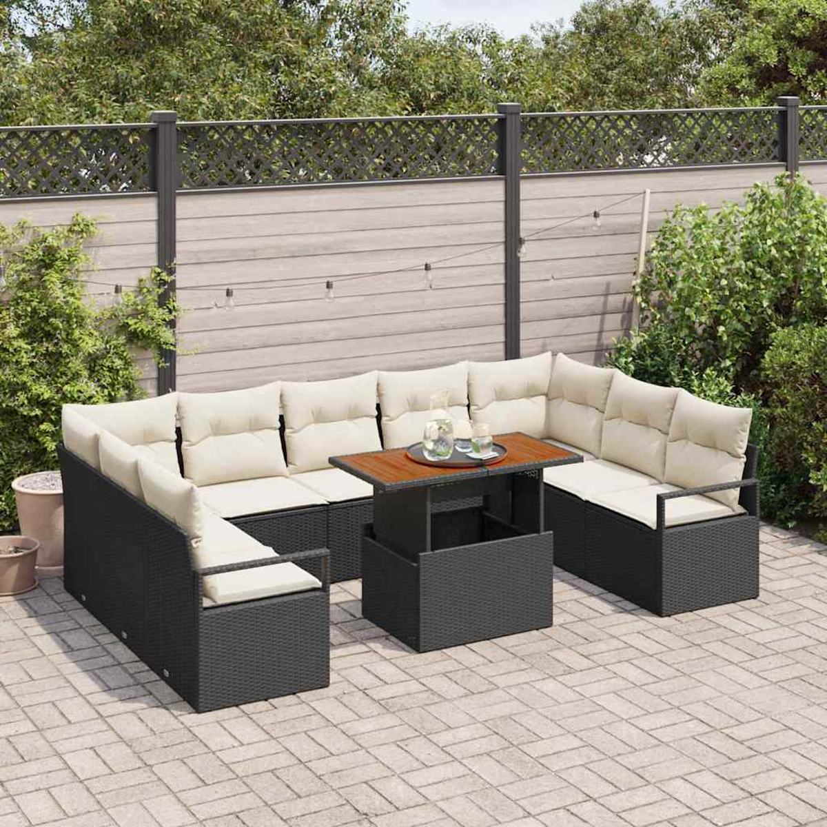 Garten-Terrassenset mit schwarzer Korbmöbel, beigen Kissen und einem Holztisch. Im Hintergrund ein Zaun und Grün.