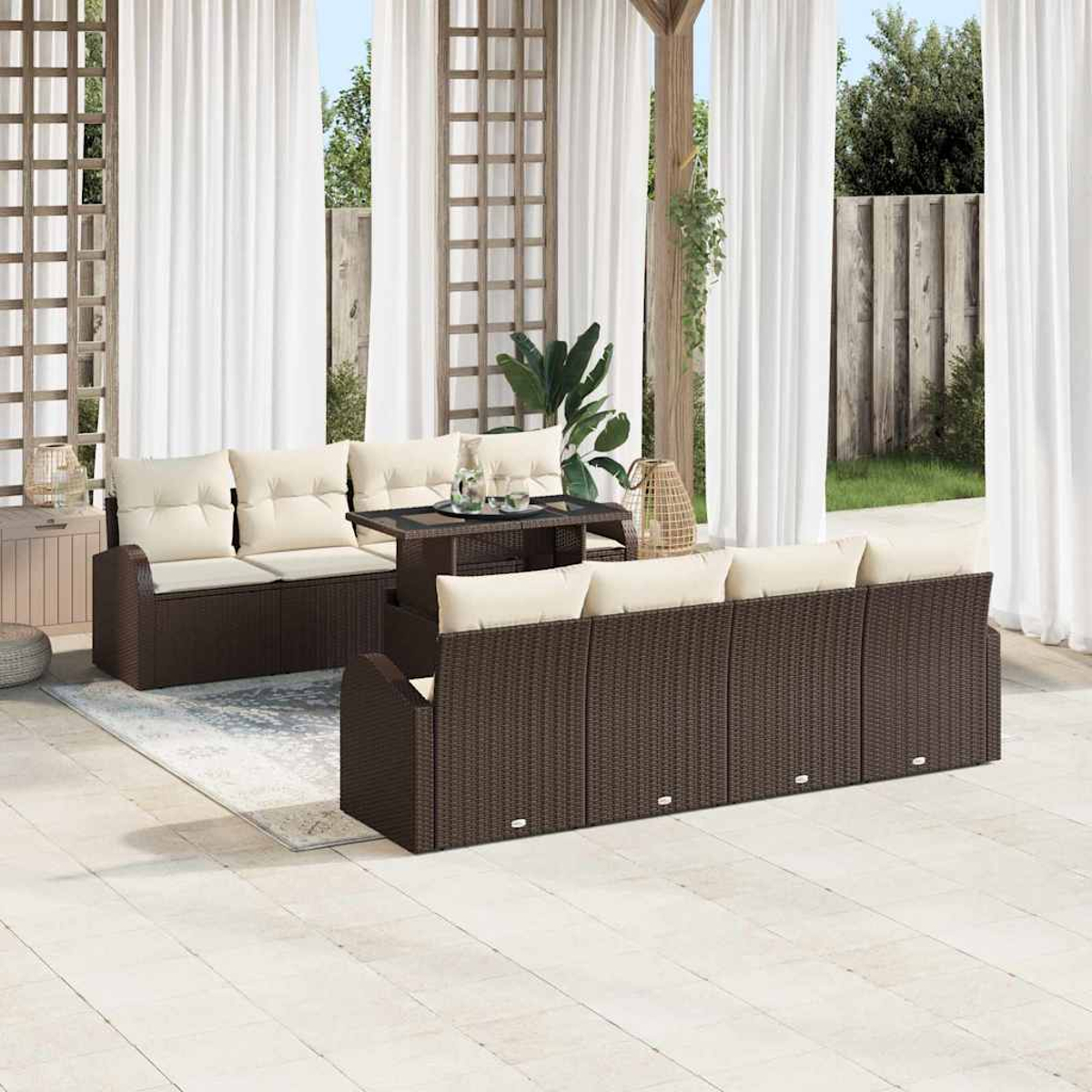 Gartenmöbel-Set mit braunen Korbs Sofas, cremefarbenen Kissen und einem Tisch mit Glasplatte. Weiße Vorhänge und grüne Pflanzen sichtbar.