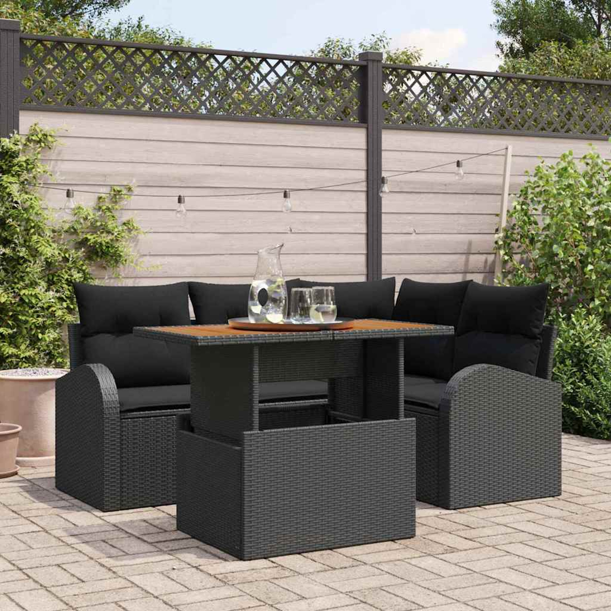 Juego de patio al aire libre con cojines negros, una mesa de madera y dos sillas. Una jarra y vasos están sobre la mesa. Una valla de madera al fondo.