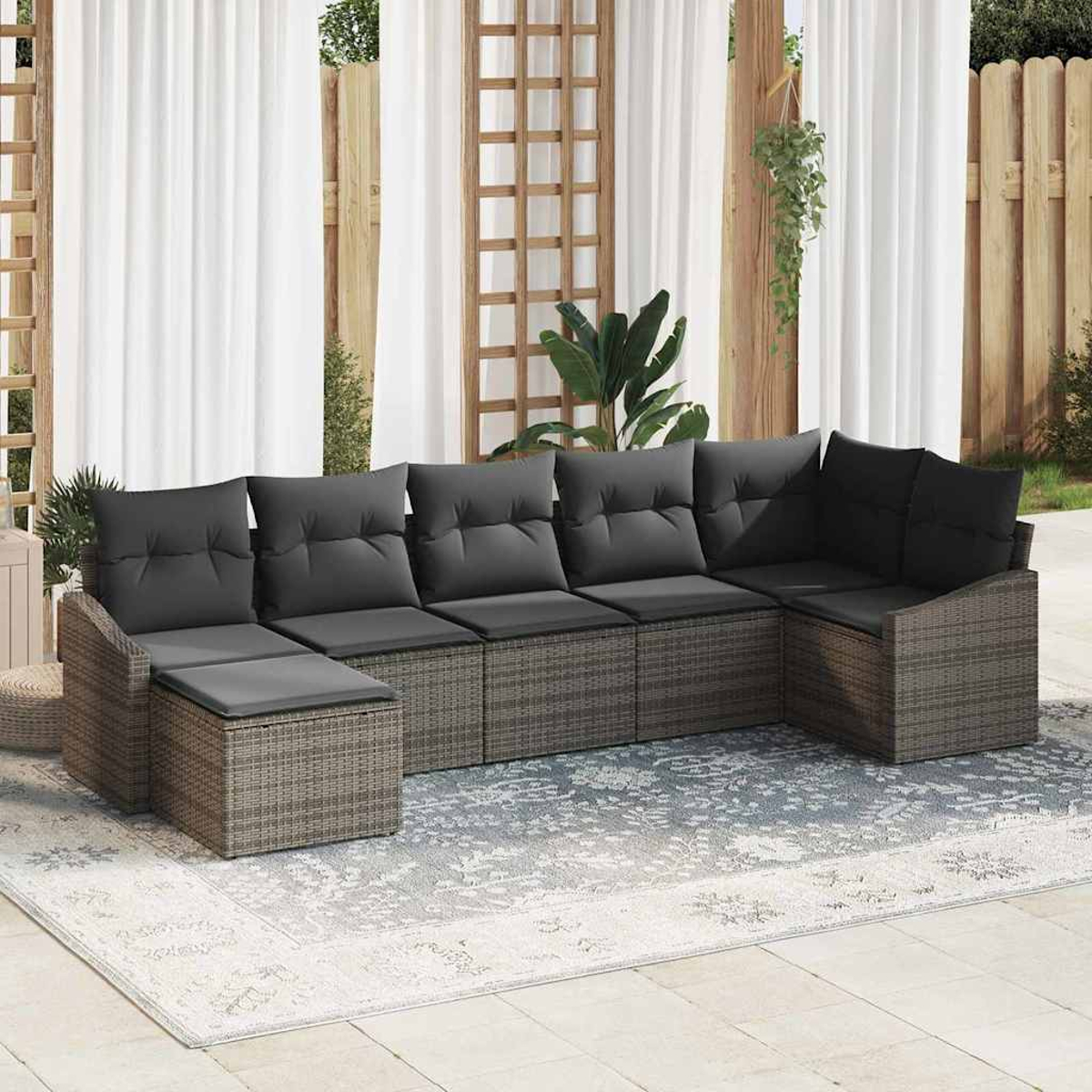 Geflochtenes Ecksofa mit grauen Kissen auf einem gemusterten Teppich, auf einer Terrasse. Weiße Vorhänge und Holzzaun im Hintergrund.