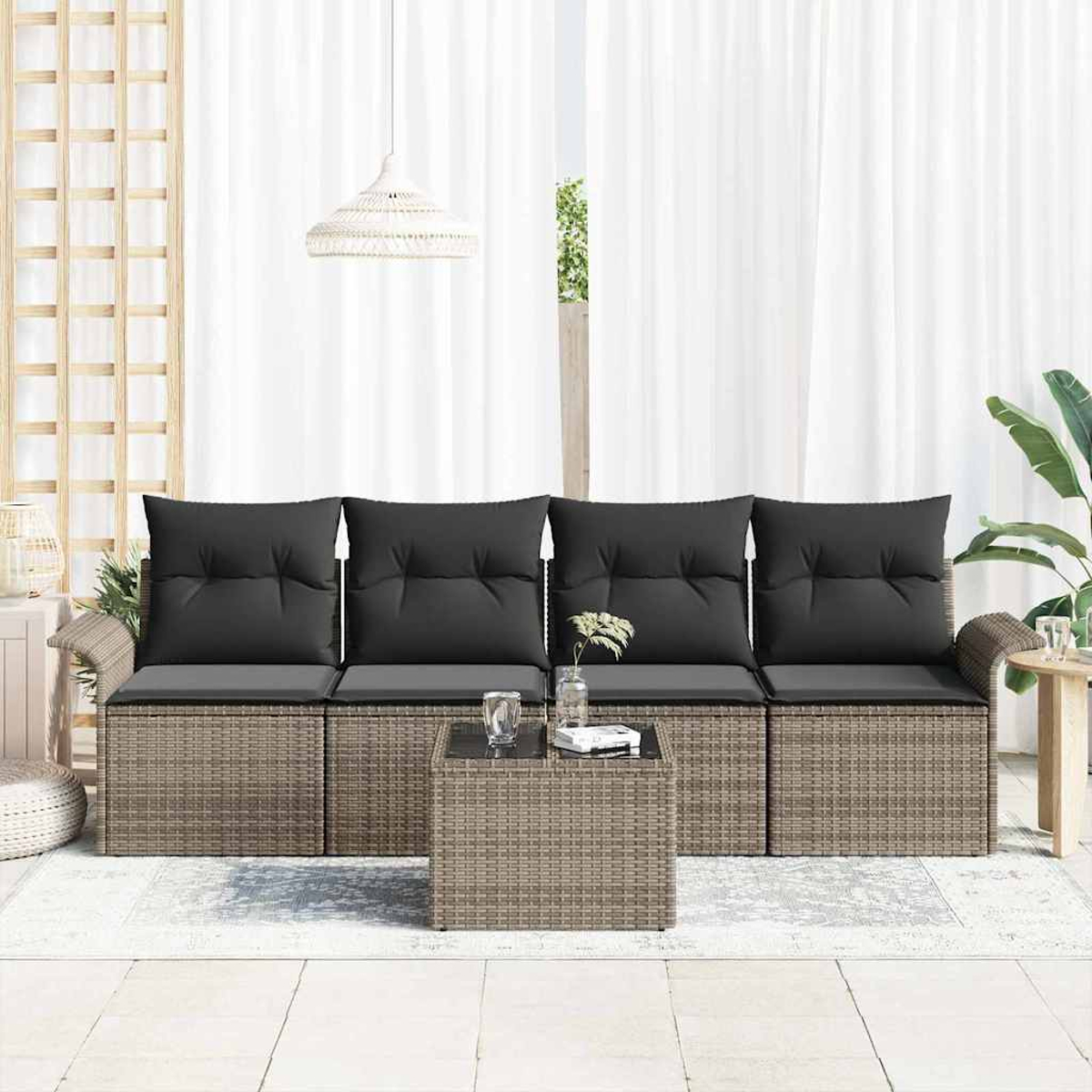 Ein Rattan-Außensofa mit schwarzen Kissen steht vor weißen Vorhängen. Ein kleiner Tisch und eine Pflanze befinden sich rechts.