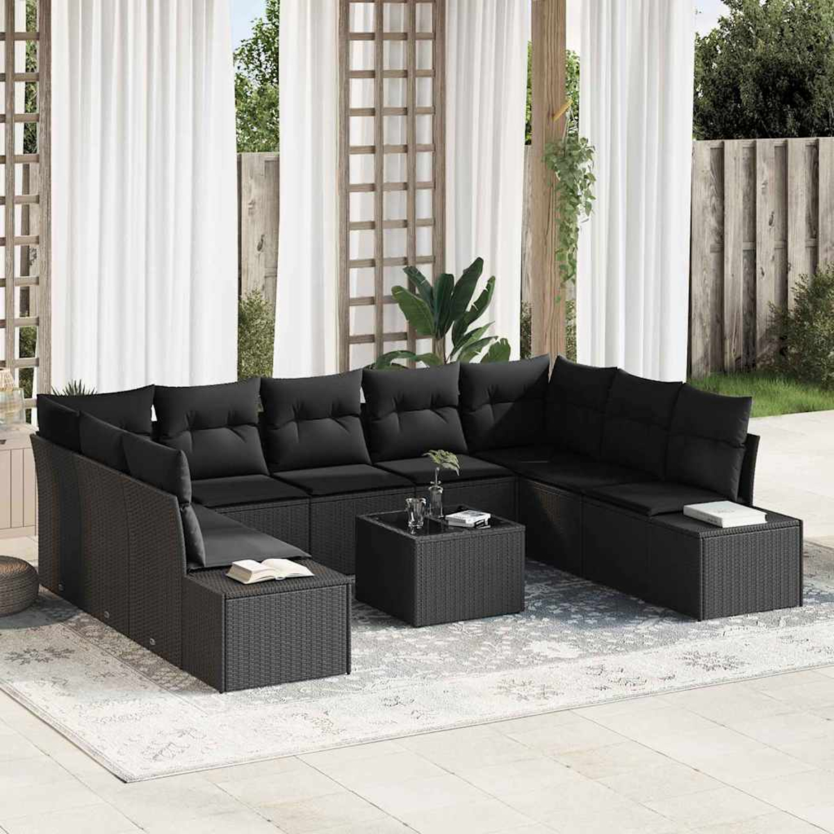 Schwarzes Outdoor-Rattan-Ecksofa mit schwarzen Kissen und einem kleinen quadratischen Tisch auf einem gemusterten Teppich. Weiße Vorhänge im Hintergrund.