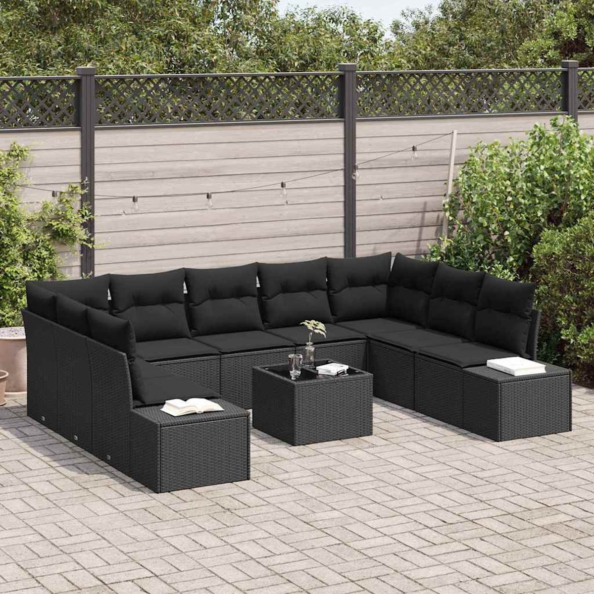 Schwarzes Rattan-Ecksofa mit Kissen auf einer gepflasterten Terrasse. Ein kleiner Tisch mit Gläsern und einem Buch ist vorhanden. Zaun und Pflanzen im Hintergrund.