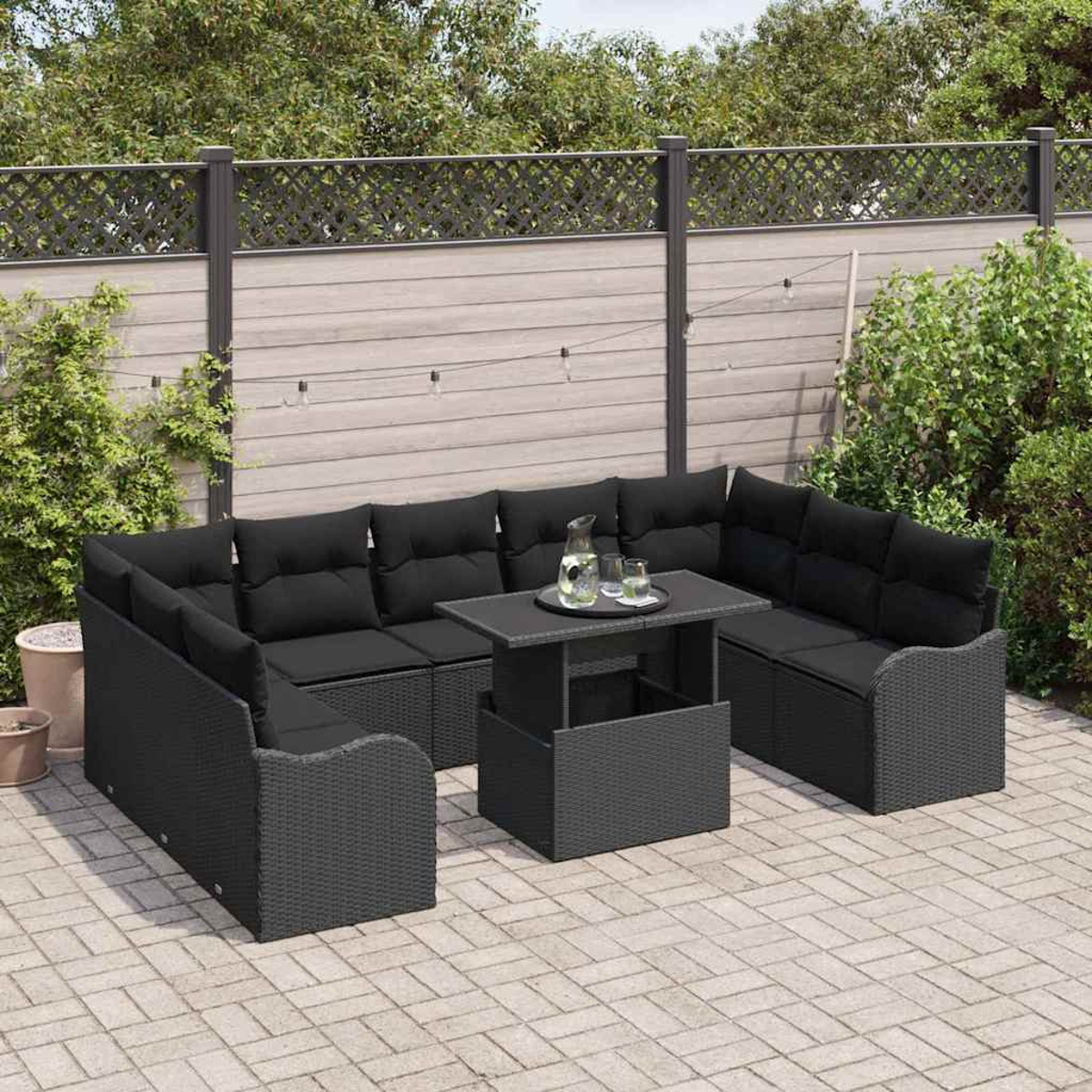 Garten-Terrassenset mit schwarzen Kissen. Enthält einen Tisch mit Glas und Wasserkrug. Umgeben von einem Holzzaun und Grün.