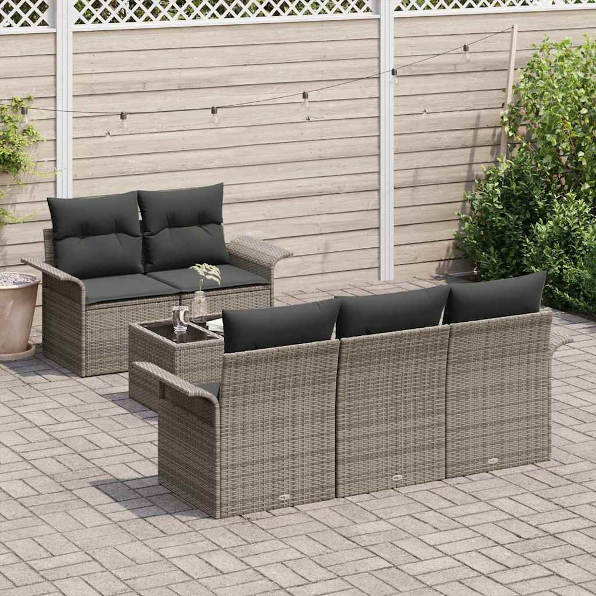 Gartenmöbel-Set aus Rattan auf einer Terrasse. Es umfasst ein Sofa, eine Doppelsitz und einen Tisch mit schwarzen Kissen, vor einem Holzzaun.