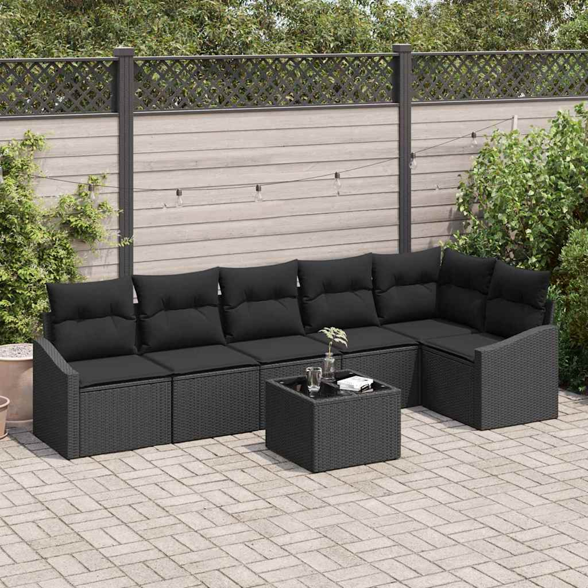 Gartenmöbel-Set. Dunkelgraues Korbmöbel mit schwarzen Kissen, kleinem Tisch und Ziegelpflaster. Hintergrund: Zaun und Pflanzen.