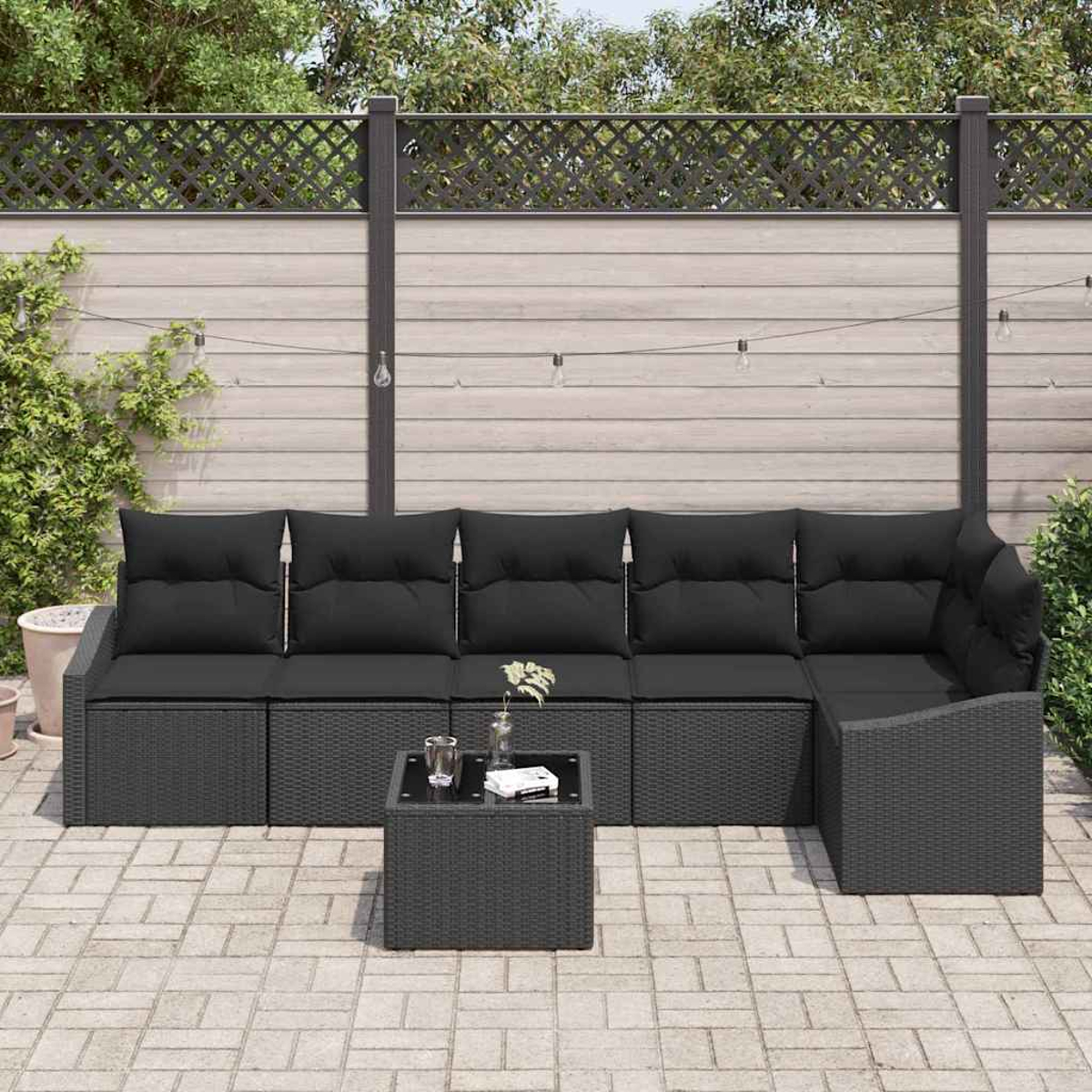 Outdoor-Ecksofa mit schwarzen Kissen und einem kleinen Tisch. Auf einer Backstein-Terrasse mit Holzzaun und dekorativen Lichtern.