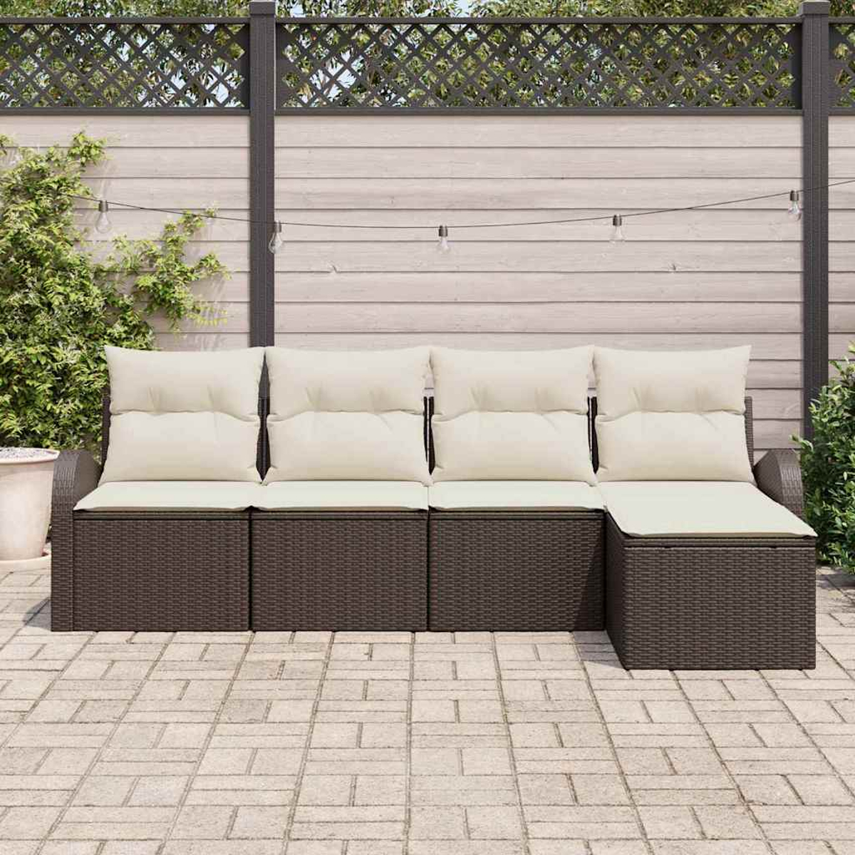 Ein braunes Outdoor-Ecksofa aus Korbgeflecht mit cremefarbenen Kissen steht auf einer Terrasse. Ein Holzzaun und Grün bilden den Hintergrund.
