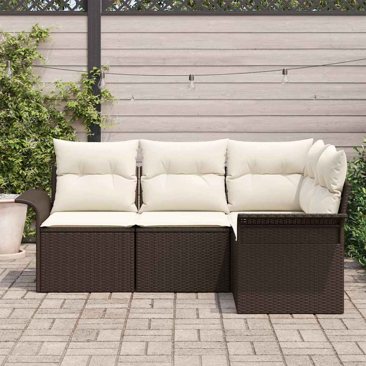 Ein braunes Rattan-Ecksofa mit cremefarbenen Kissen steht auf einer Backsteinterrasse. Dahinter ist ein Holzzaun mit Lichterketten.