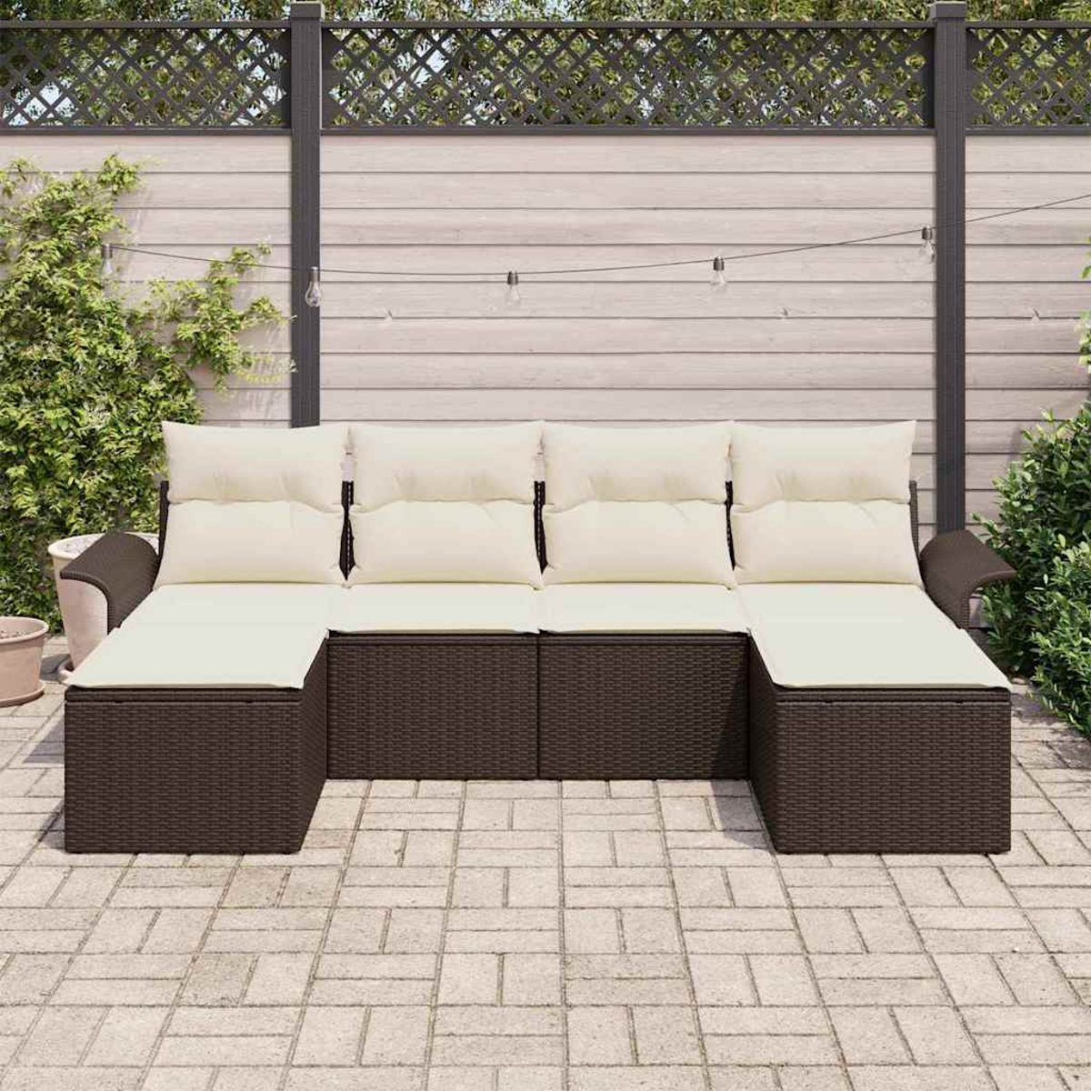 Braunes Korbsofa mit cremefarbenen Kissen auf einer Terrasse. Hintergrund: Zaun, grüne Pflanzen und Lichterketten.