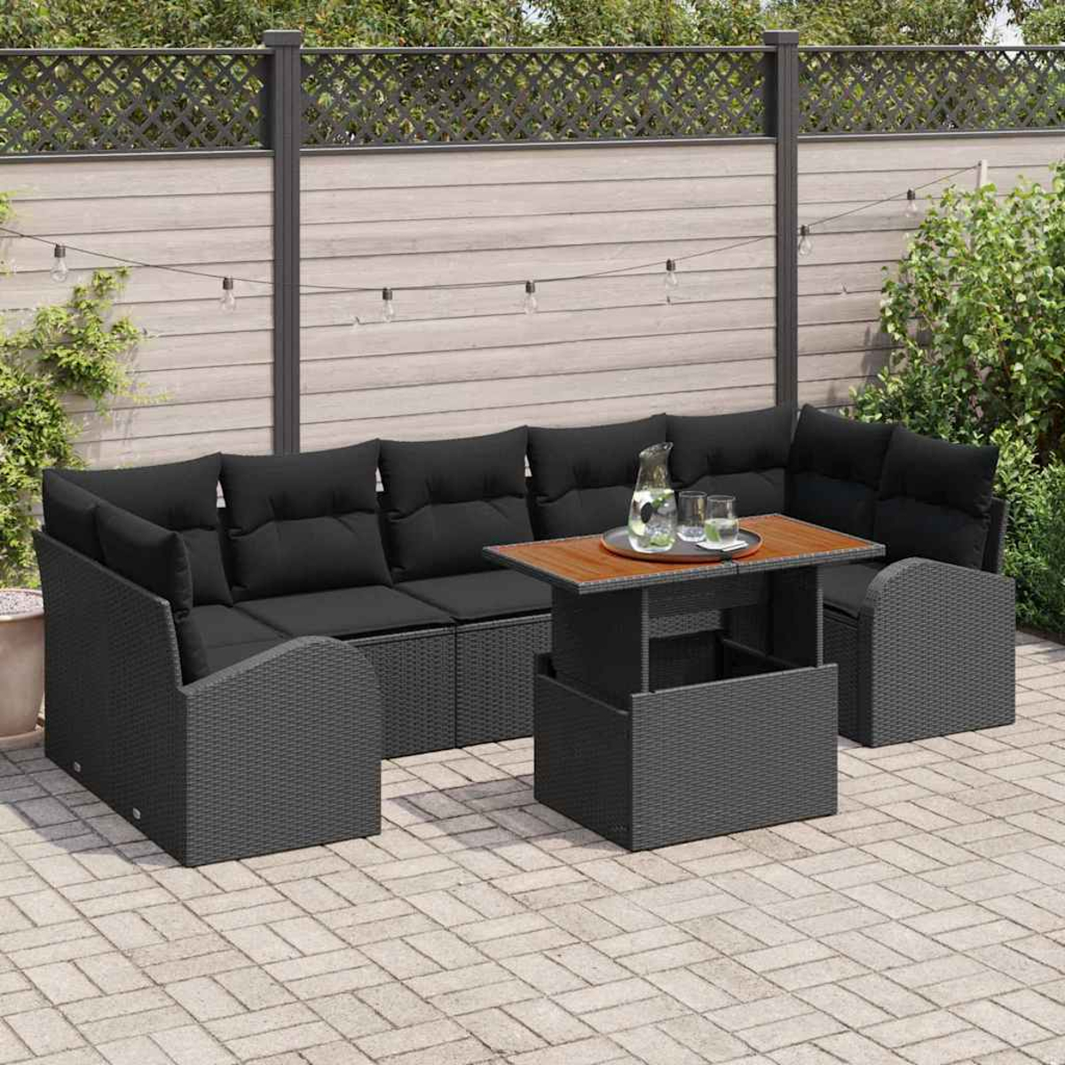 Terrassenset im Freien mit schwarzen Kissen und einem Holztisch. Auf dem Tisch stehen eine Kanne und Gläser. Im Hintergrund ein Zaun und Grün.