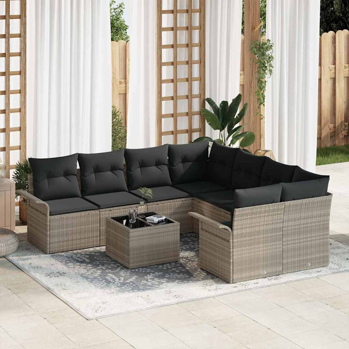 Ein Sitzbereich im Freien mit Rattanmöbeln, schwarzen Kissen, einem Couchtisch und einem gemusterten Teppich. Weiße Vorhänge und ein Holzzaun.