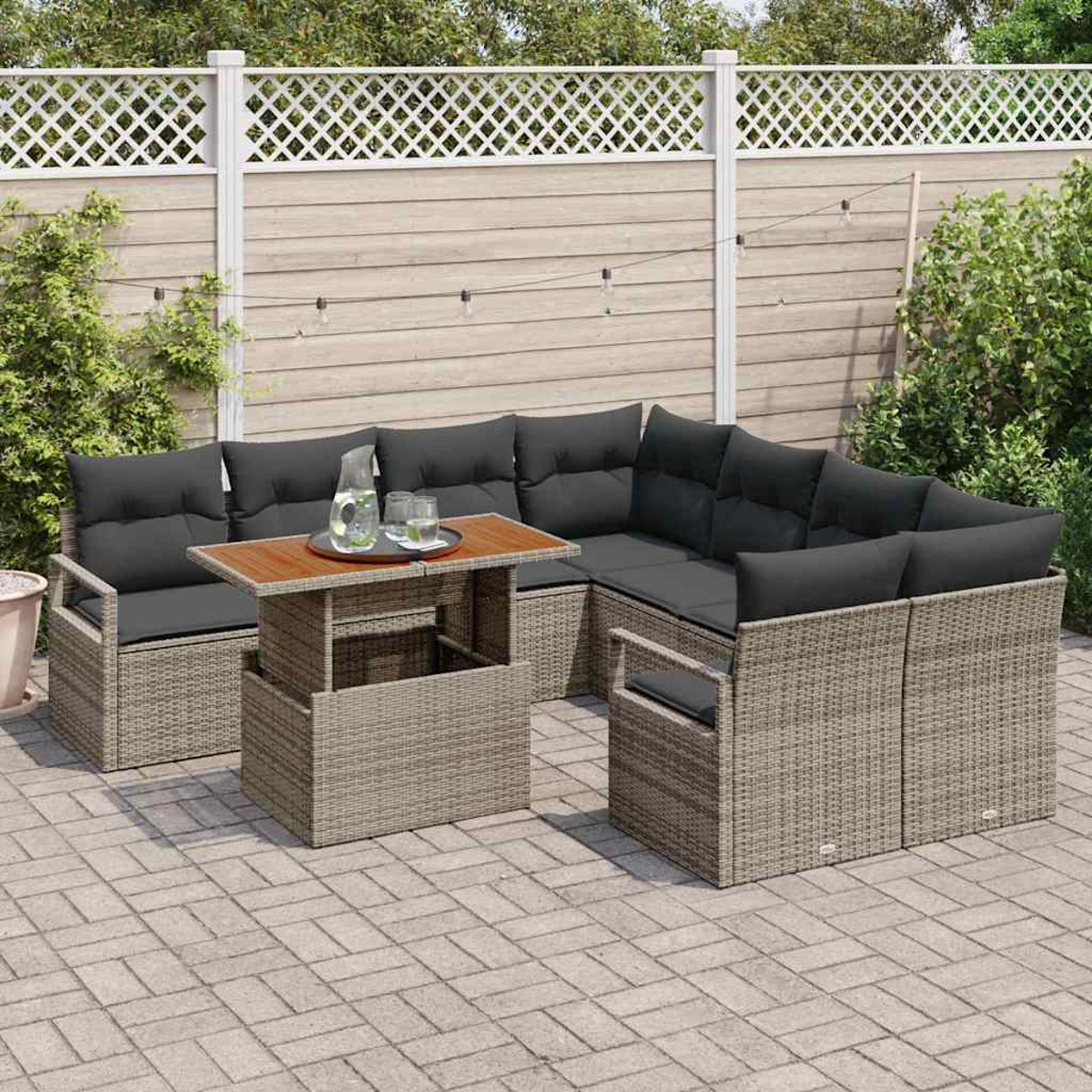 Ein graues Ecksofa aus Korbgeflecht mit schwarzen Kissen steht auf einer Terrasse. Ein Holztisch hält Getränke. Ein weißer Zaun und Grünzeug dahinter.