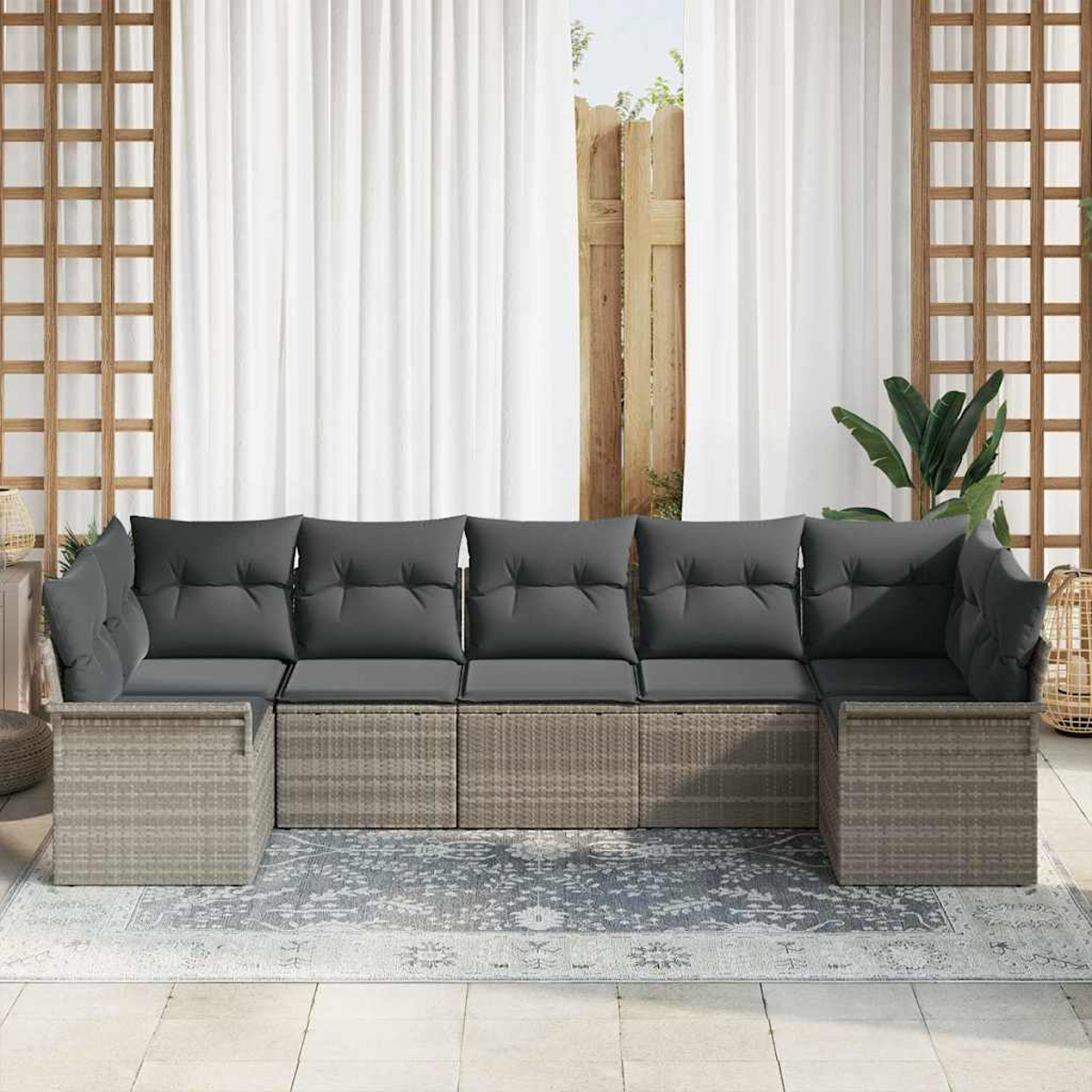 Ein graues Ecksofa mit Kissen steht auf einem gemusterten Teppich. Weiße Vorhänge und Holzgitter befinden sich hinter dem Sofa.