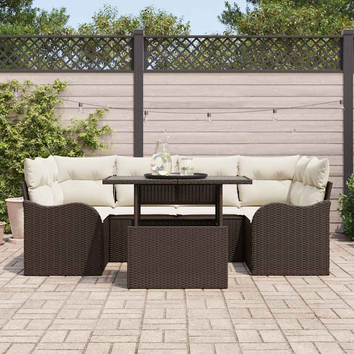 Terrassenmöbel-Set im Freien mit dunkelbraunen Korbmöbeln, cremefarbenen Kissen und einem kleinen Tisch. Hintergrund ist ein Holzzaun und Grün.