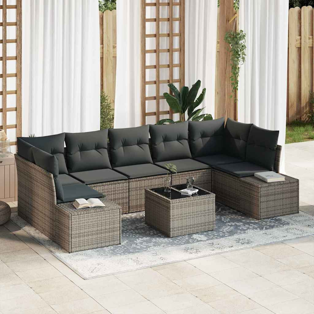 Outdoor-Ecksofa mit grauen Kissen und einem kleinen Tisch auf einem gemusterten Teppich, vor weißen Vorhängen und Holzzaun.
