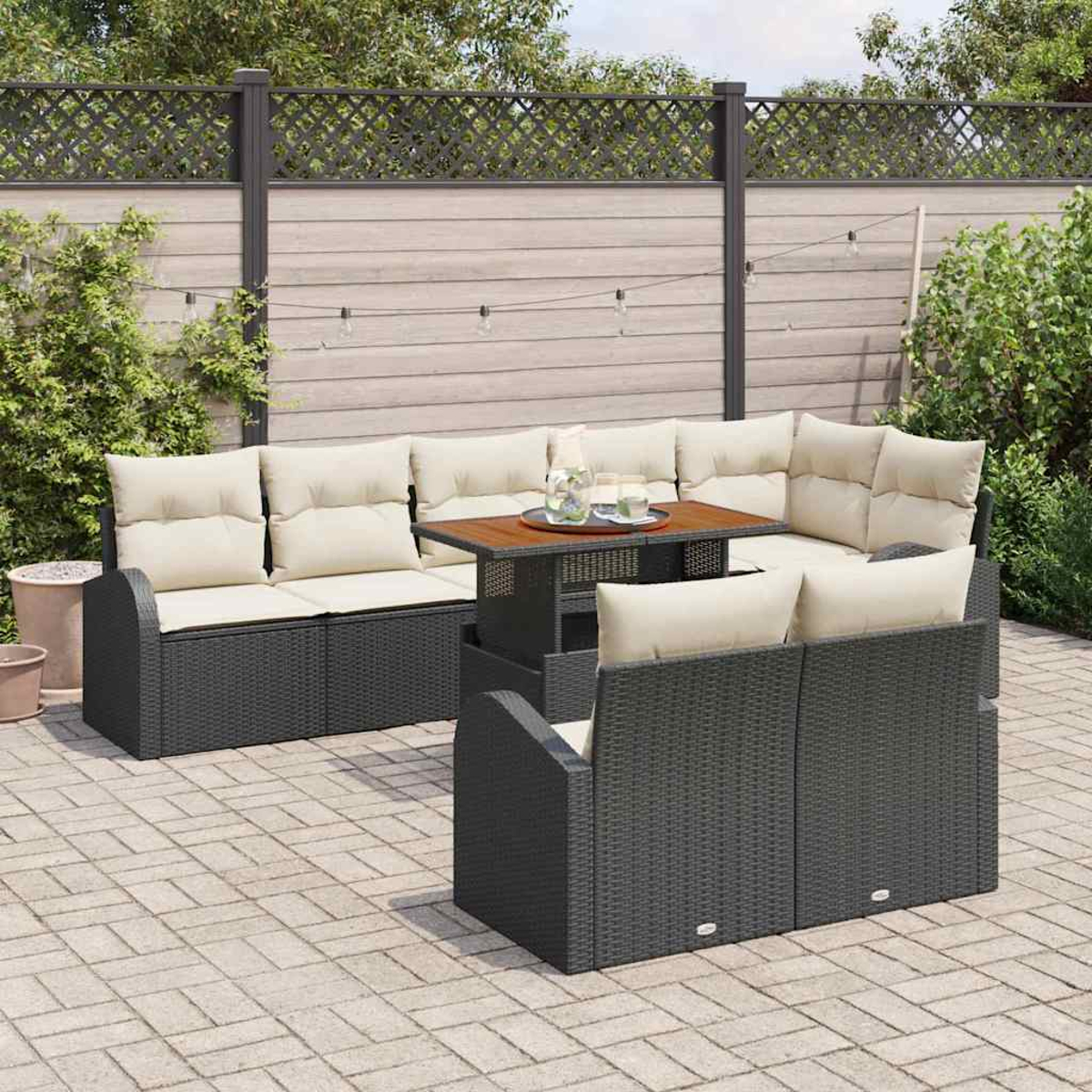Terrassenset mit dunklen Möbeln im Wicker-Stil und cremefarbenen Kissen. Ein Holztisch steht im Zentrum. Ein Zaun und Pflanzen sind im Hintergrund.
