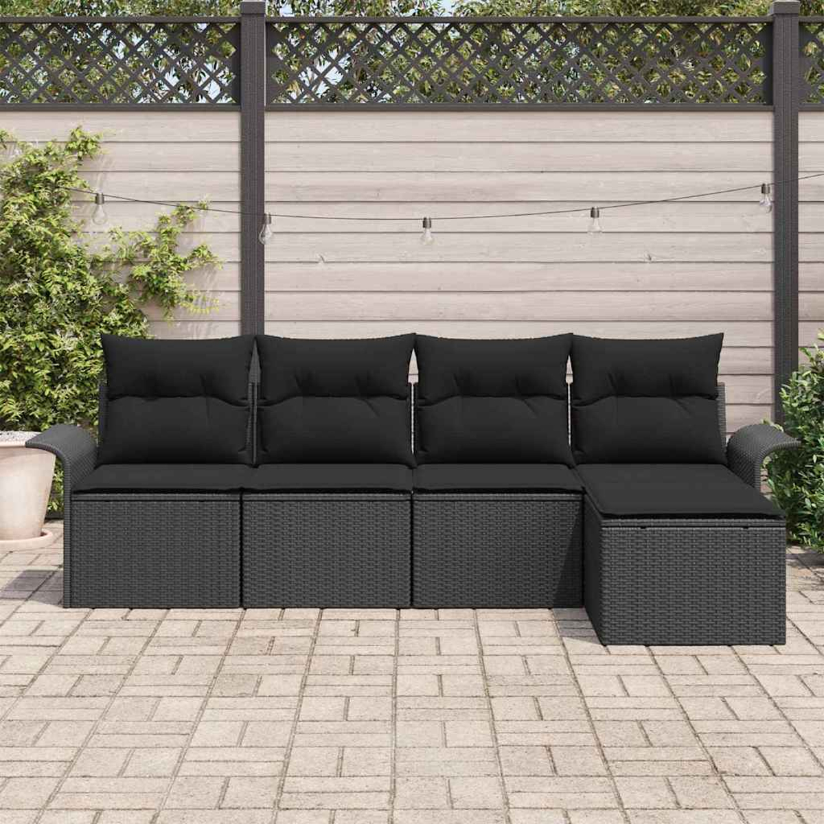 Ein schwarzes Outdoor-Ecksofa aus Korbgeflecht mit schwarzen Kissen steht auf einer Ziegelterrasse. Im Hintergrund befindet sich ein Zaun mit Lichterketten.