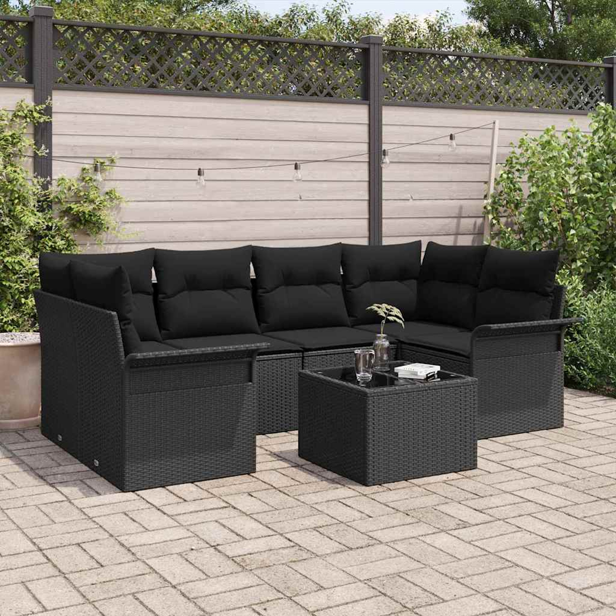 Schwarzes Outdoor-Ecksofa-Set aus Korbgeflecht mit schwarzen Kissen und einem kleinen Tisch auf einer Ziegelterrasse. Im Hintergrund sind ein Zaun und Grün.