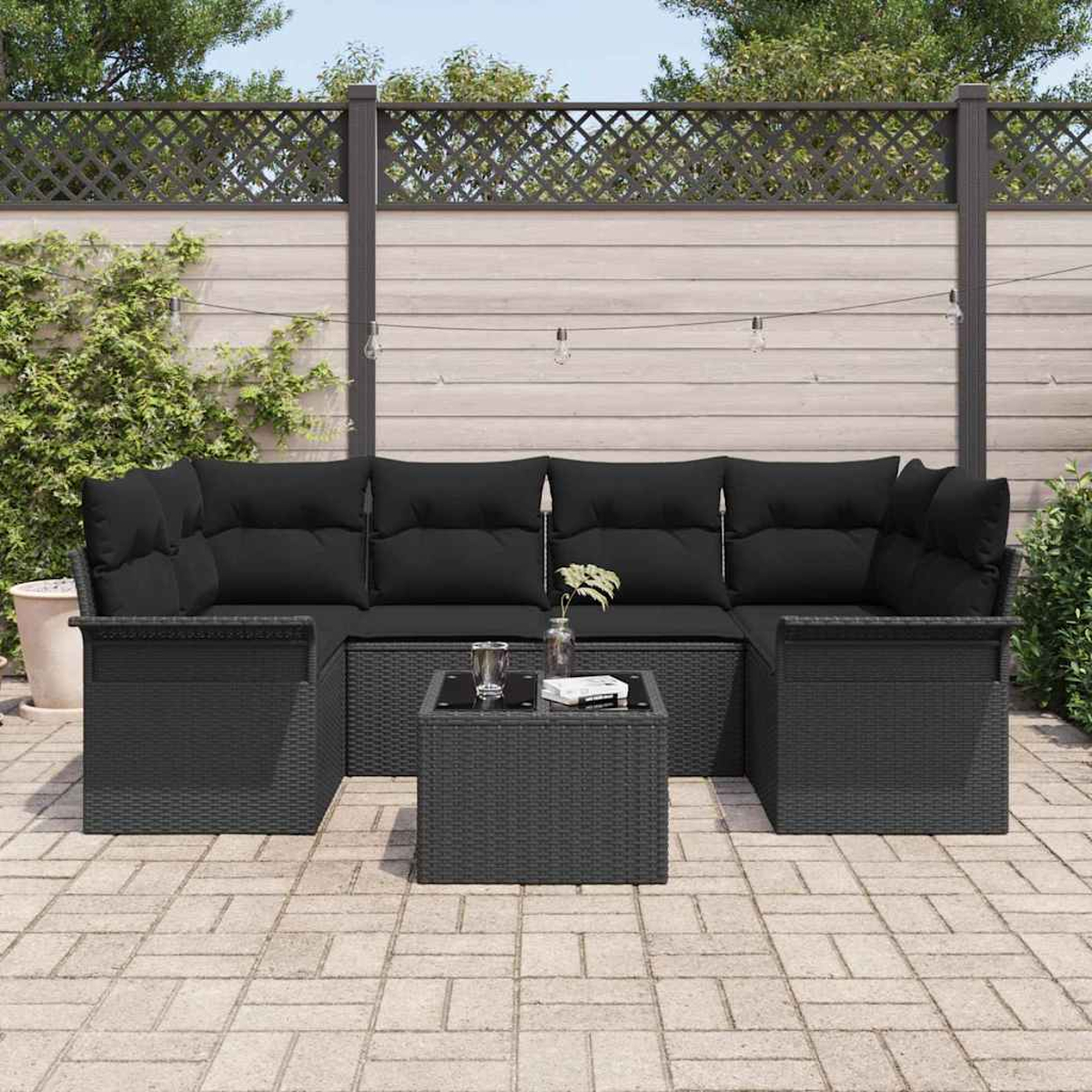 Schwarzes Outdoor-Korbsessel-Set mit Kissen, kleinem Tisch und einem Glas Wasser. Das Set steht auf einer Ziegelterrasse mit Zaun.