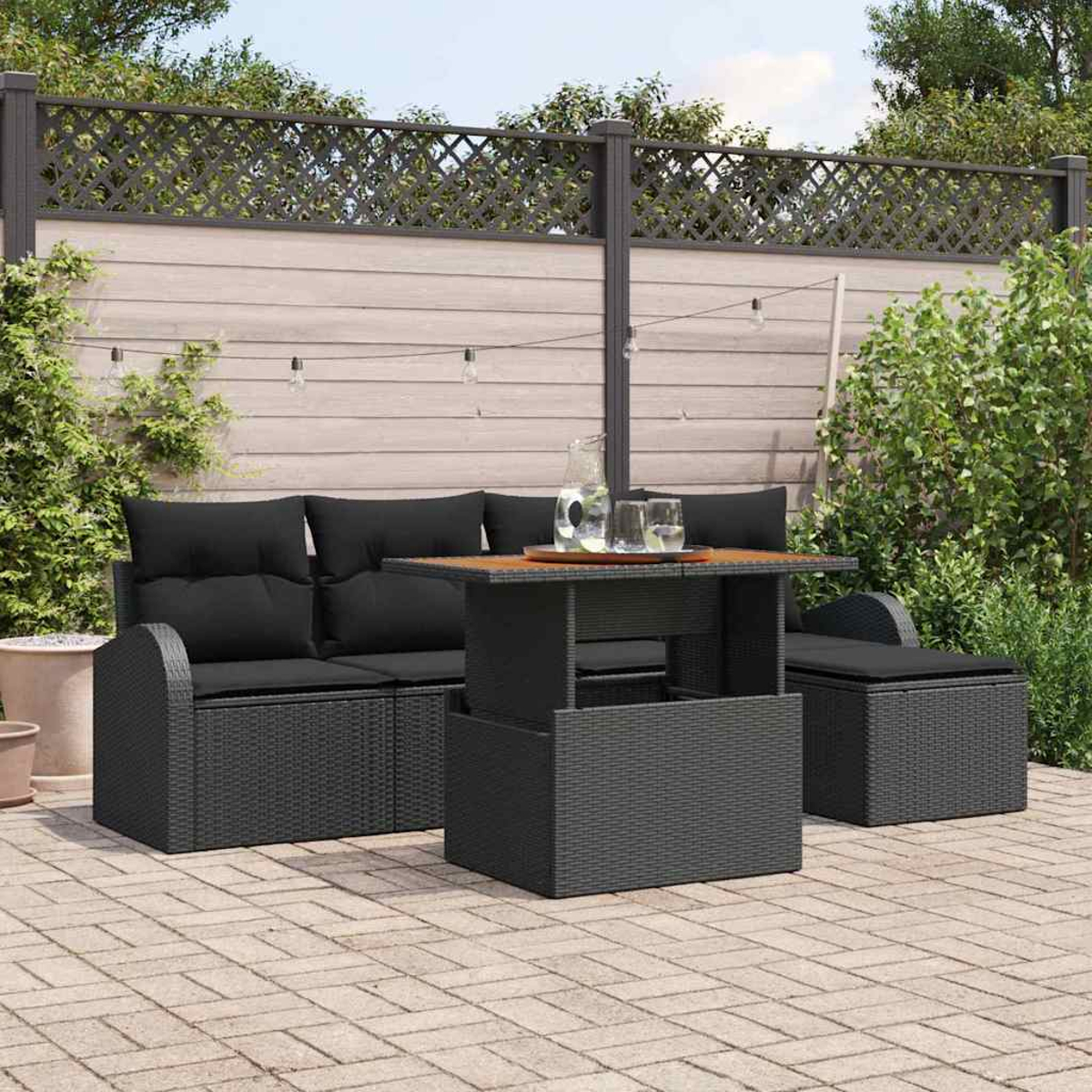 Garten-Set mit schwarzen Kissen und Tisch. Enthält Sofa, Tisch und Ottomane auf einer Ziegelterrasse mit Zaun im Hintergrund.
