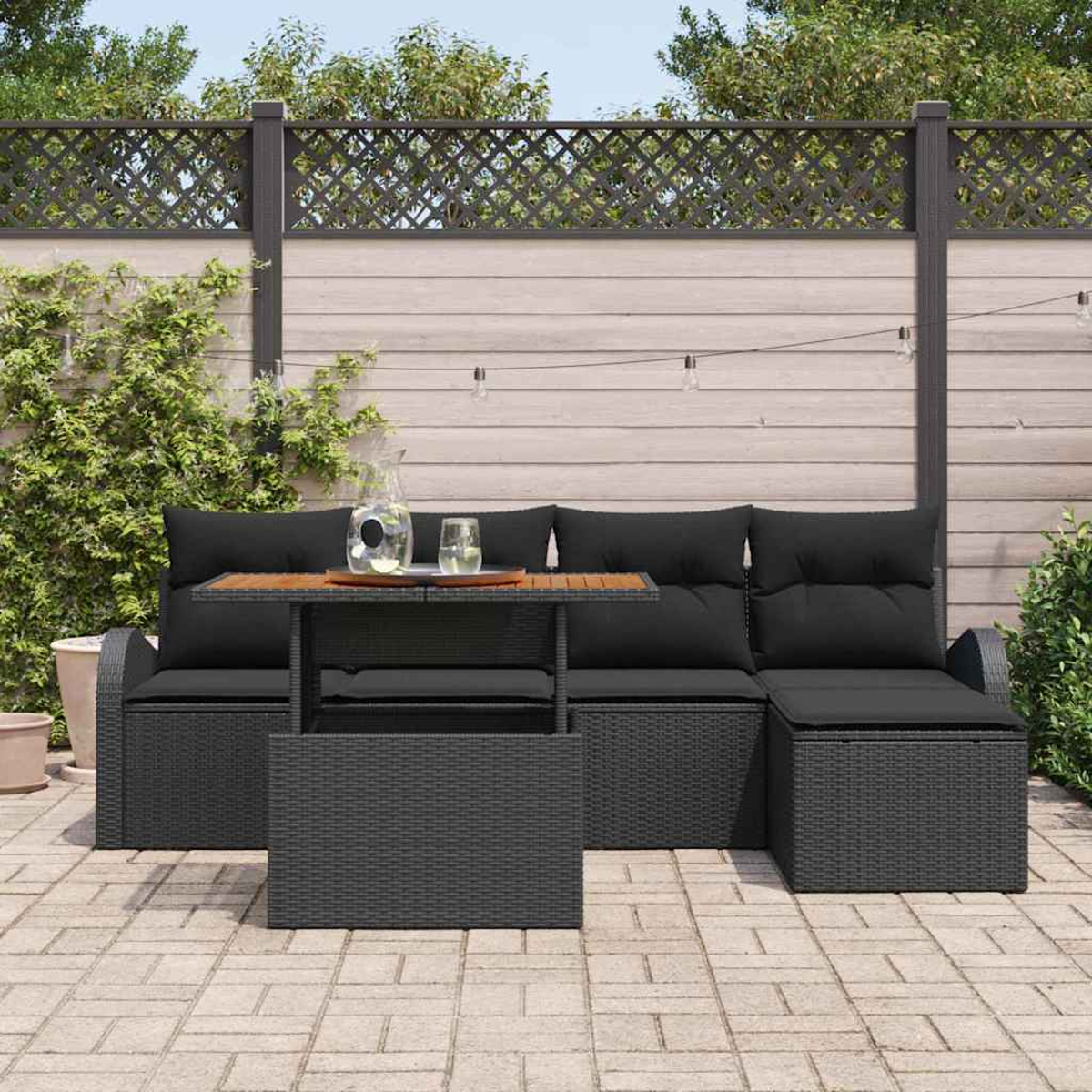 Terrassengarnitur mit schwarzen Kissen, einem Tisch mit Getränken und einem Holzzaun im Hintergrund. Gepflasterter Boden.