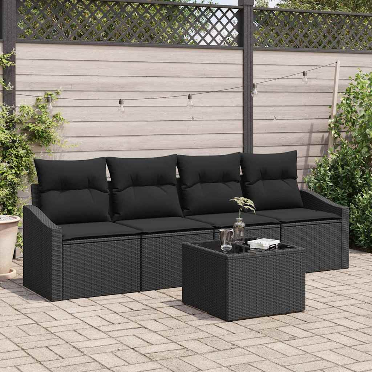 Szene im Freien mit einem schwarzen Korbsessel, Kissen und einem quadratischen Tisch. Ein Holzzaun mit Lichtern hängt über dem Sofa.