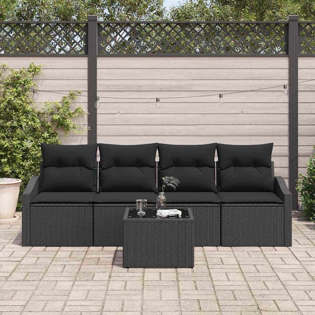Ein schwarzes Outdoor-Sofa aus Korbgeflecht mit schwarzen Kissen und einem kleinen Tisch. Im Hintergrund ein Holzzaun und Pflanzen.