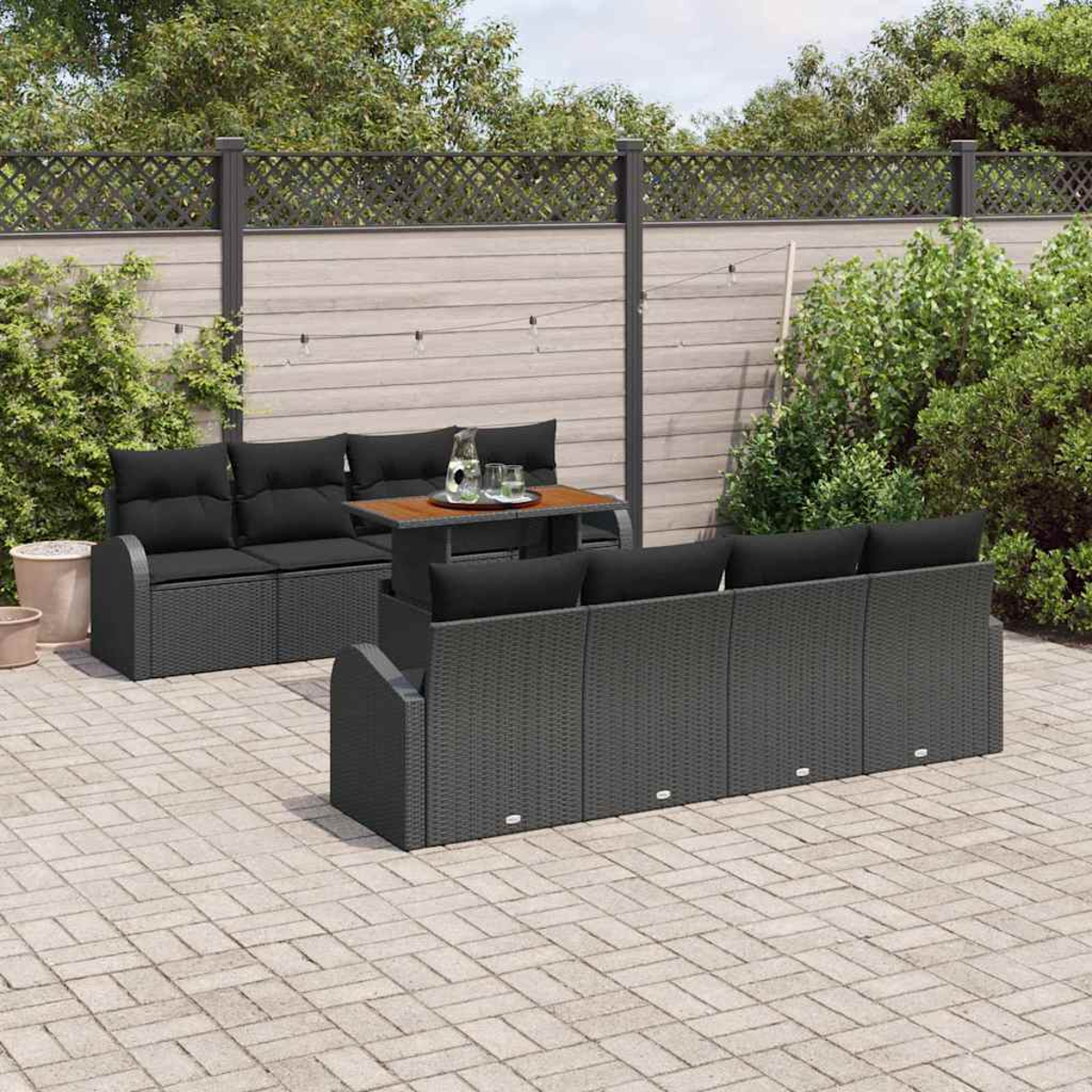Garten-Terrassenset mit schwarzen Kissen, kleinem Tisch und grauem Korbmuster vor Holzzaun und Grün.