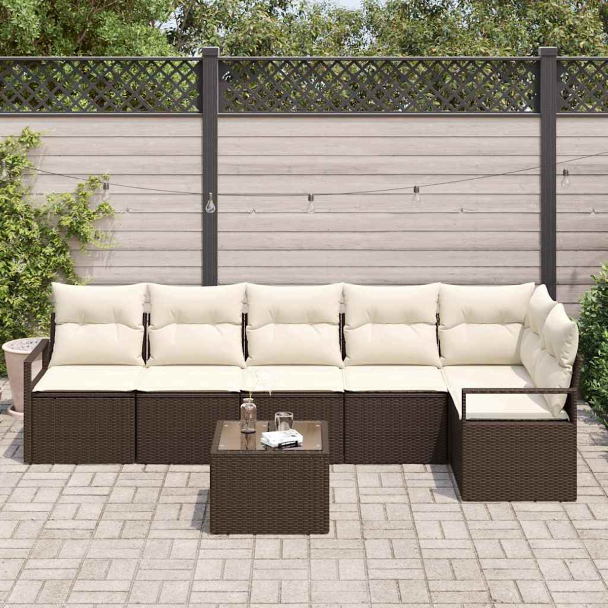 Outdoor-Ecksofa mit beigen Kissen und braunem Korbgewebe. Glastisch davor. Hintergrund: Holzzaun und Grün.