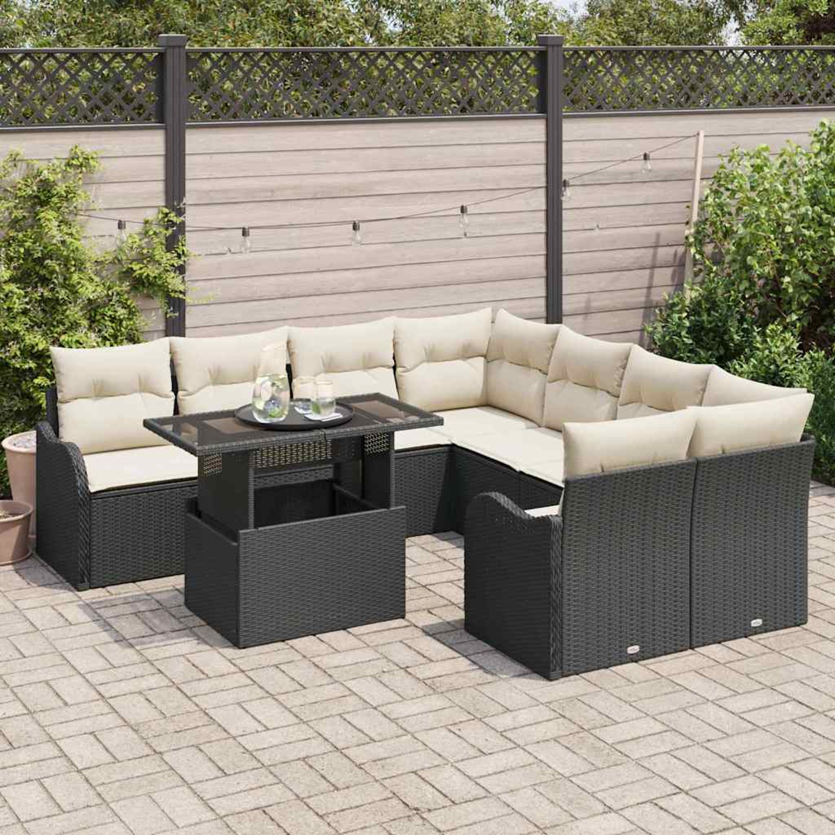 Juego de patio al aire libre con un sofá seccional de mimbre negro, cojines crema y una mesa elevable a juego, contra una valla de madera.