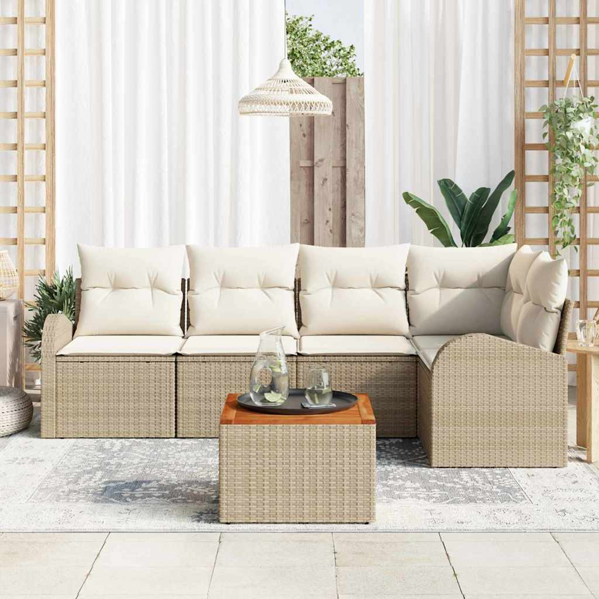 Sofá de ratán al aire libre con cojines crema, mesa de madera con bebidas y una alfombra decorativa en el patio. Cortinas blancas al fondo.