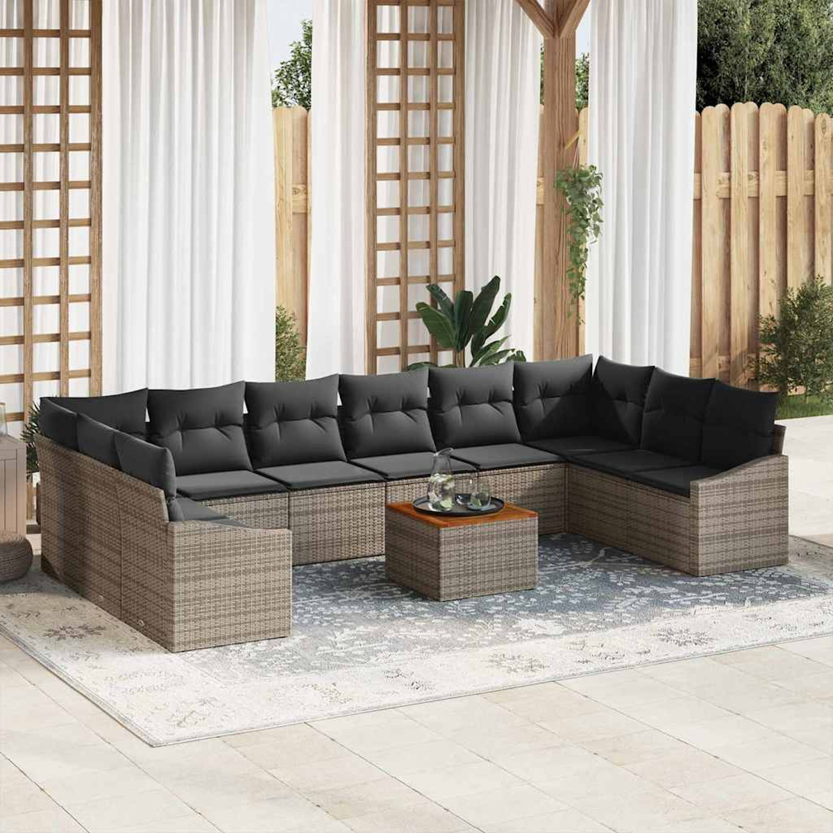 Un espacio al aire libre con sofá seccional de mimbre, cojines negros, mesa de centro de madera y una alfombra decorativa. Cortinas blancas al fondo.