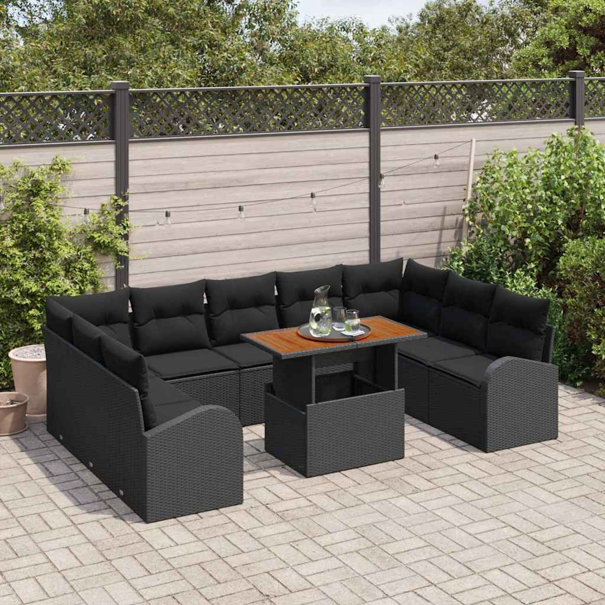 Garten-Terrassenset mit schwarzen Kissen, einem Holztisch und einer Wasserkaraffe. Auf einer gefliesten Terrasse mit Zaun und Grün.
